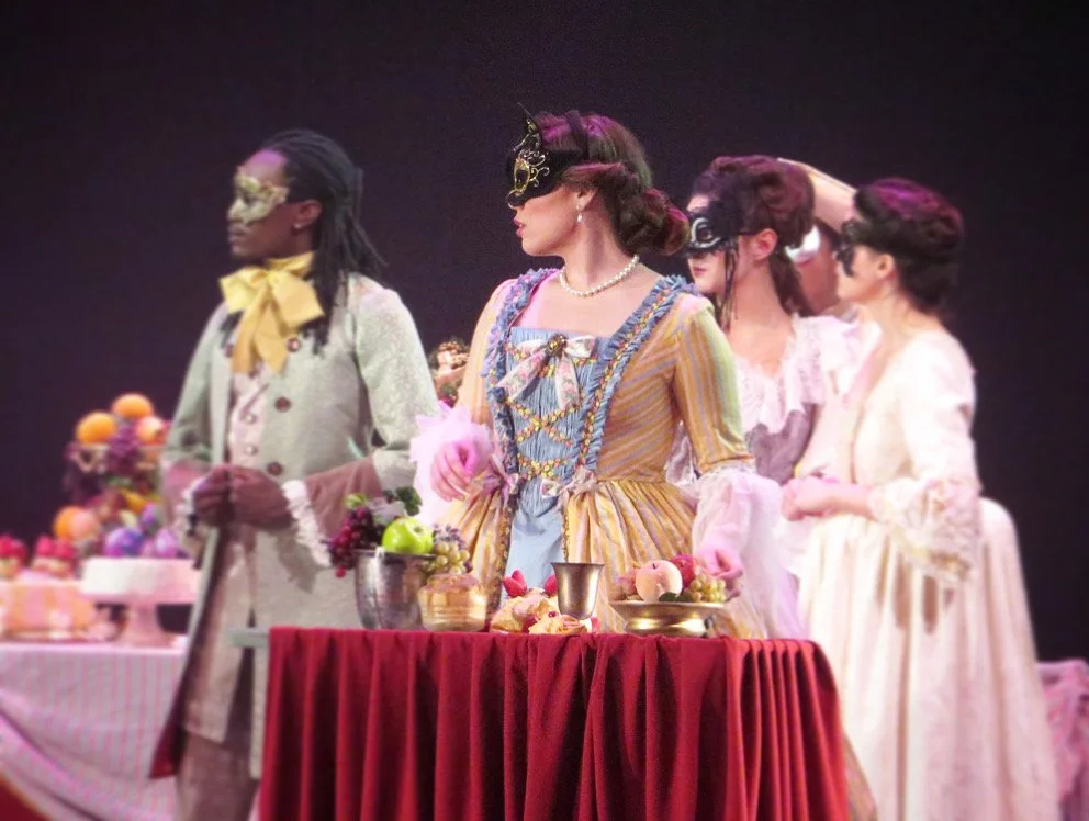 Women dressed in historical costumes wearing masks, standing around a table filled with fruits, flowers, and decorative items
