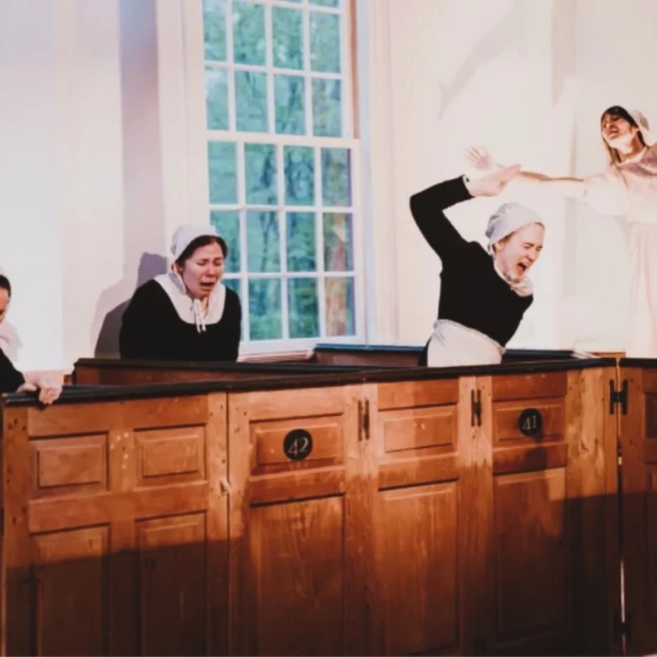 Actors dressed as maids in a theatrical play, with one acting distressed and another appearing to be in a dramatic moment, set in a room with large window and wooden paneling.