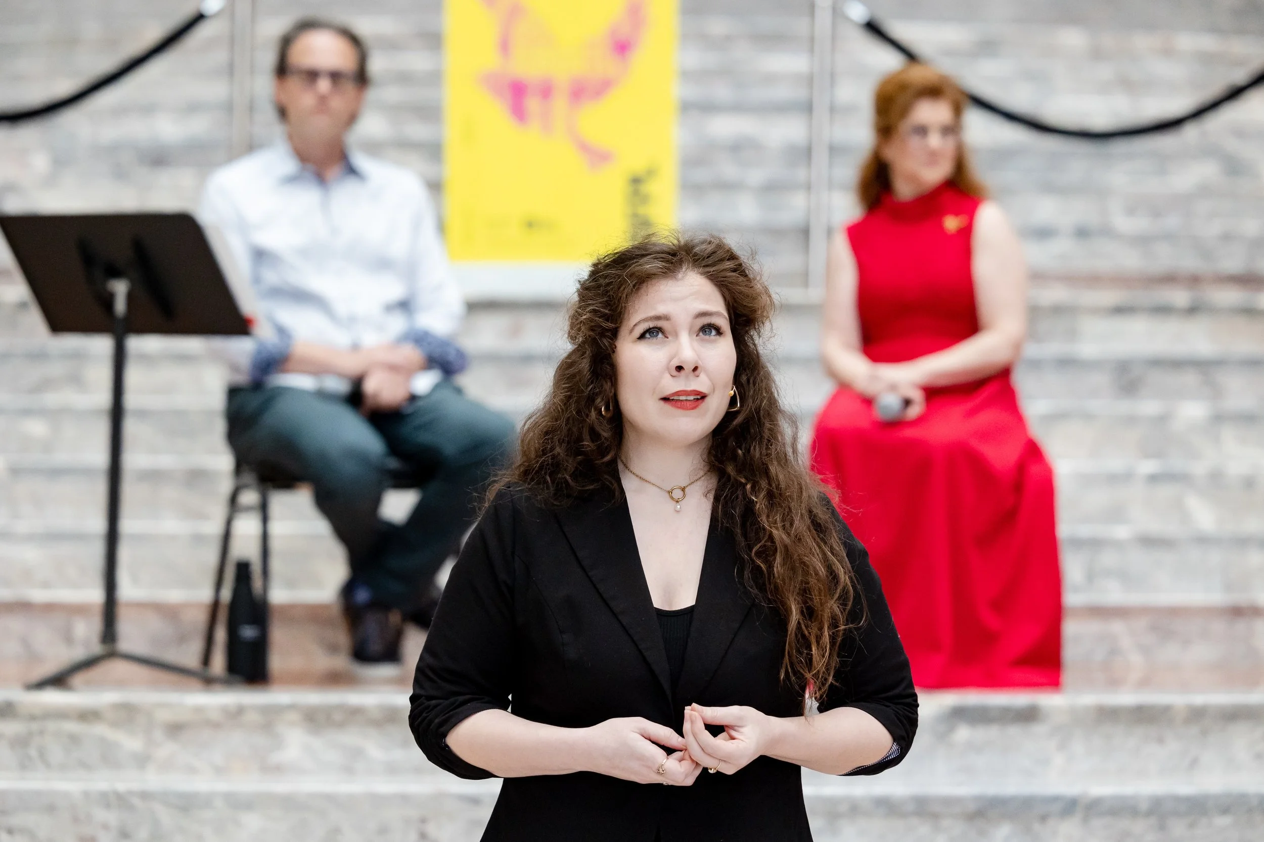 A woman with curly brown hair wearing a black blazer, sitting cross-legged with her hands clasped. Two individuals are in the background: a man in glasses and a white shirt, and a woman in a red dress holding a microphone. They are sitting on steps against a brick wall with a yellow poster behind them.