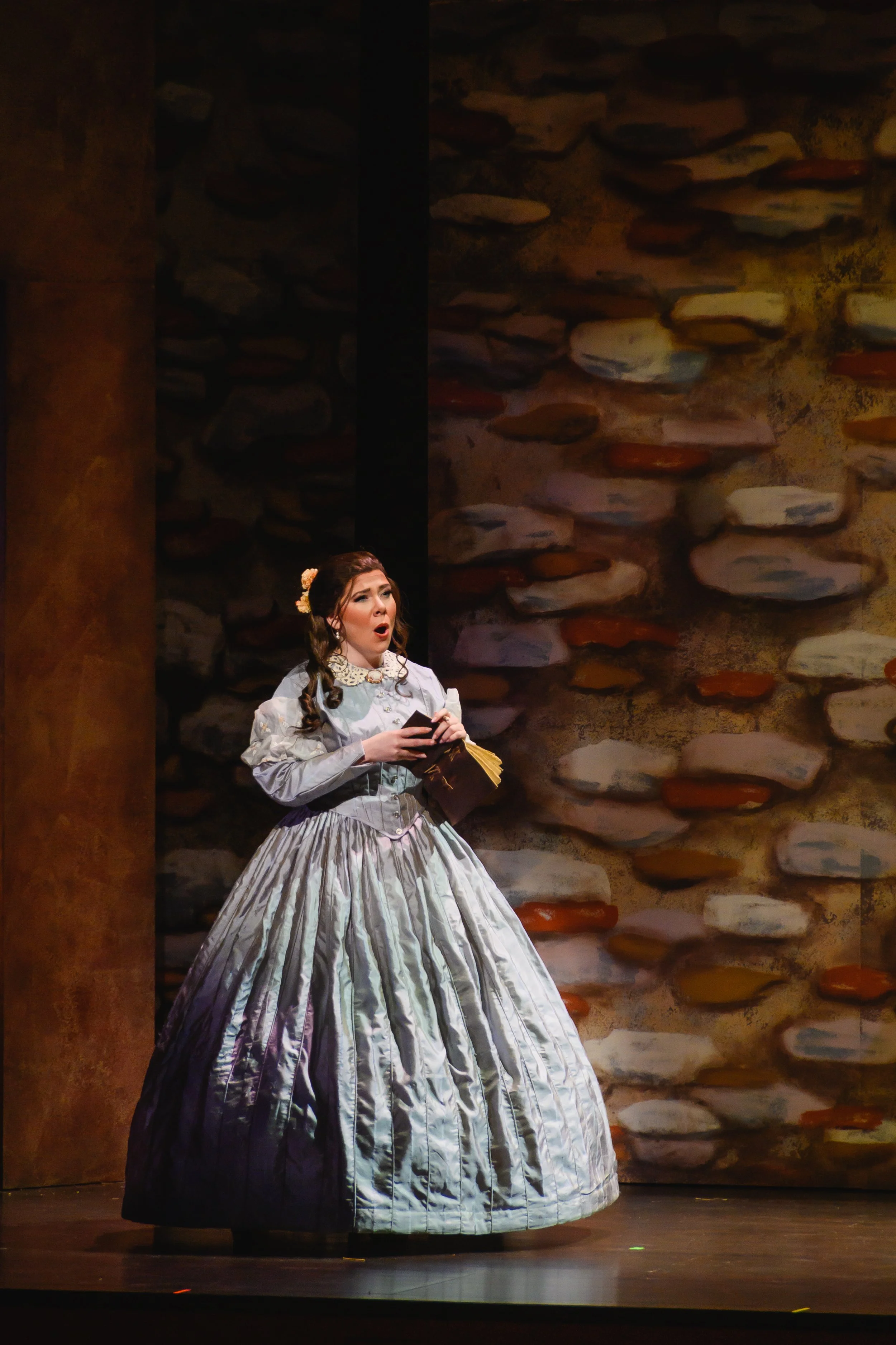 A woman in a historical costume singing on stage with a background of a stone wall.