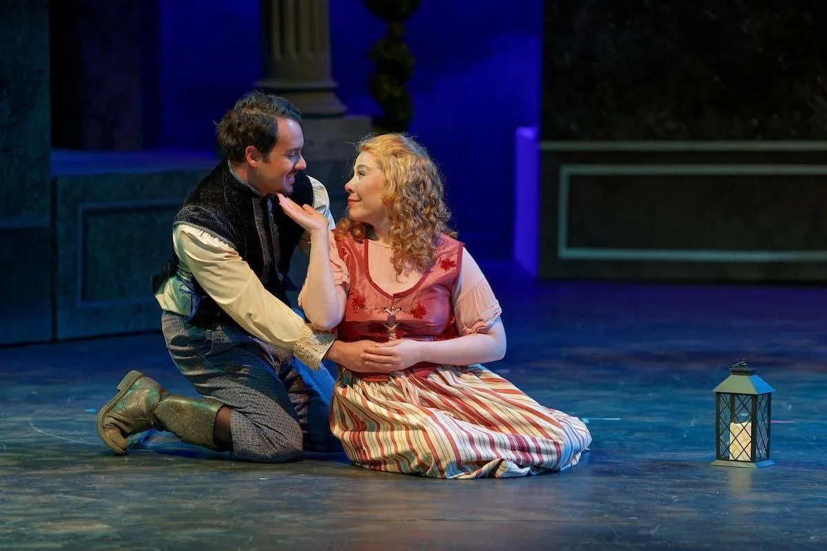 Two actors on stage performing a scene with one kneeling and the other sitting, both in period costumes, with a lantern on stage.