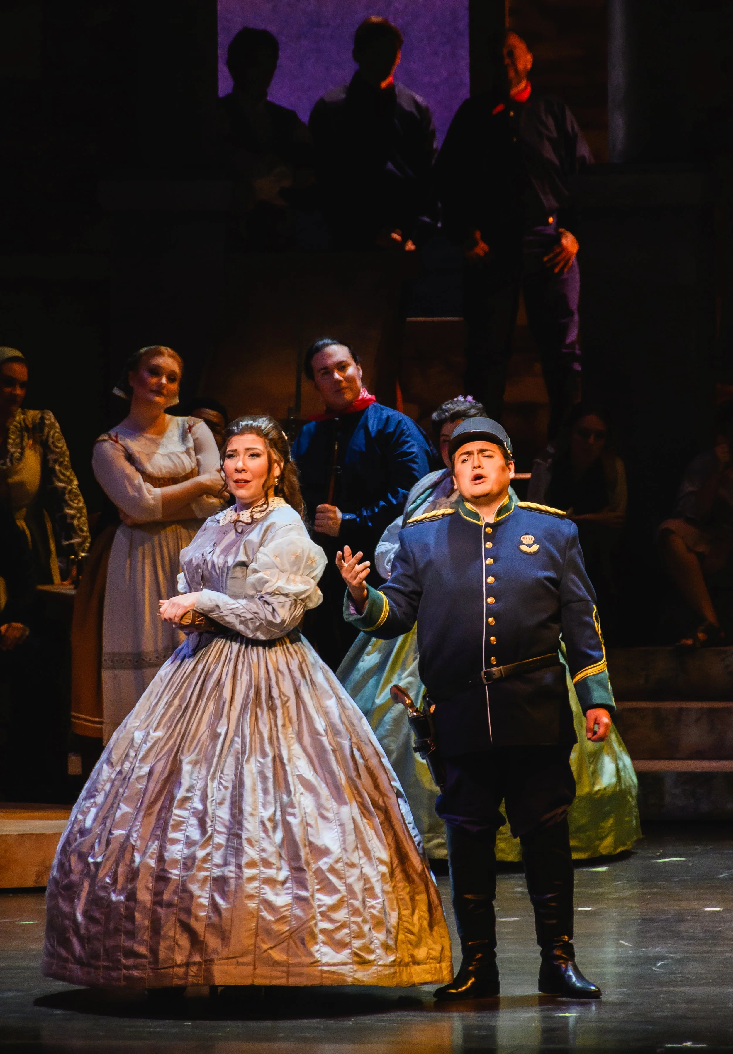Stage scene from a theatrical performance showing actors in period costumes, including a woman in a voluminous white and beige striped dress, a man in a military uniform, and others in historical attire, with a dark background and dramatic theatrical lighting.