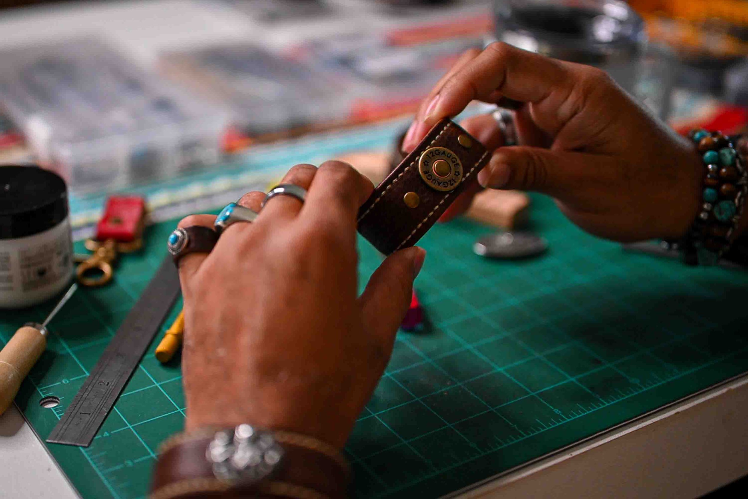 A Black leather artisan works on the handcrafted Koa cuff at a workshop table, adjusting its brass-toned hardware by hand among leatherworking tools and supplies on a green cutting mat.