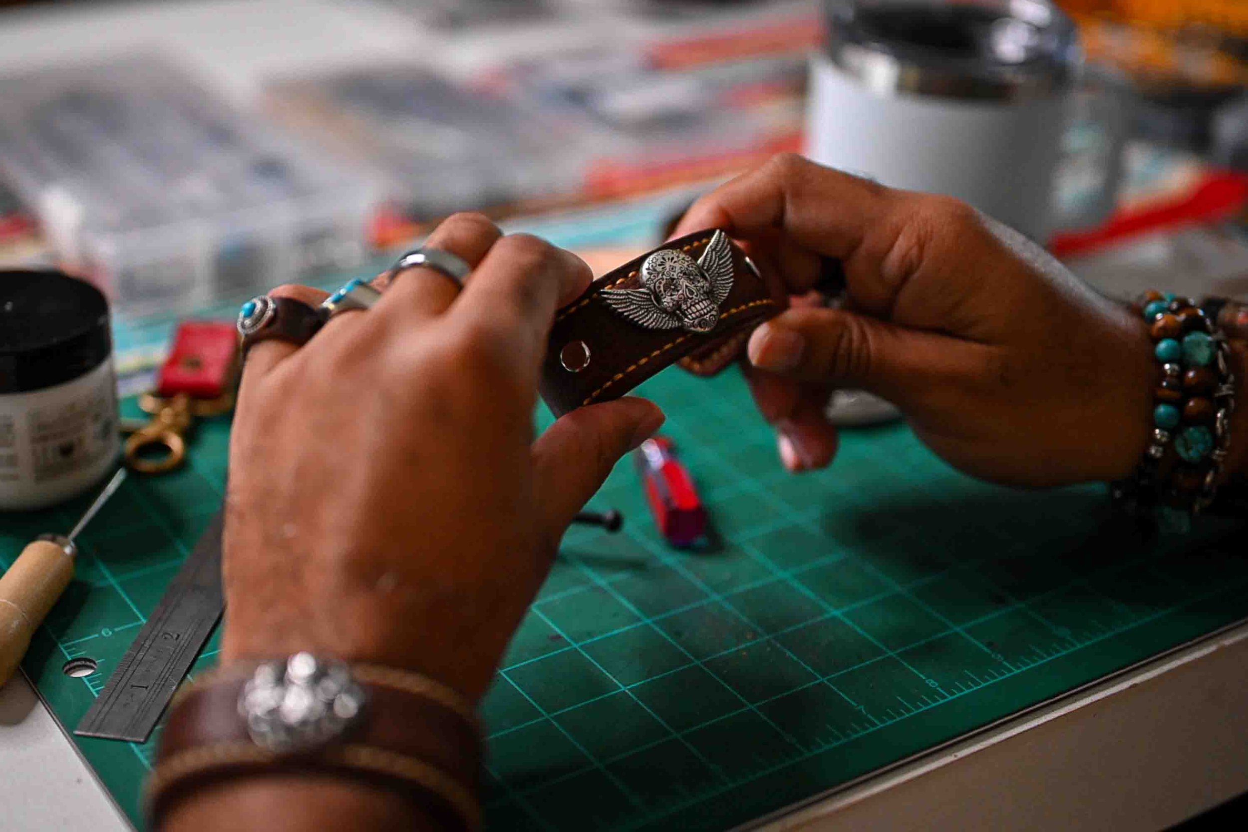 Person working on a leather bracelet with a skull and wings design, jewelry tools and a cutting mat on the workspace.