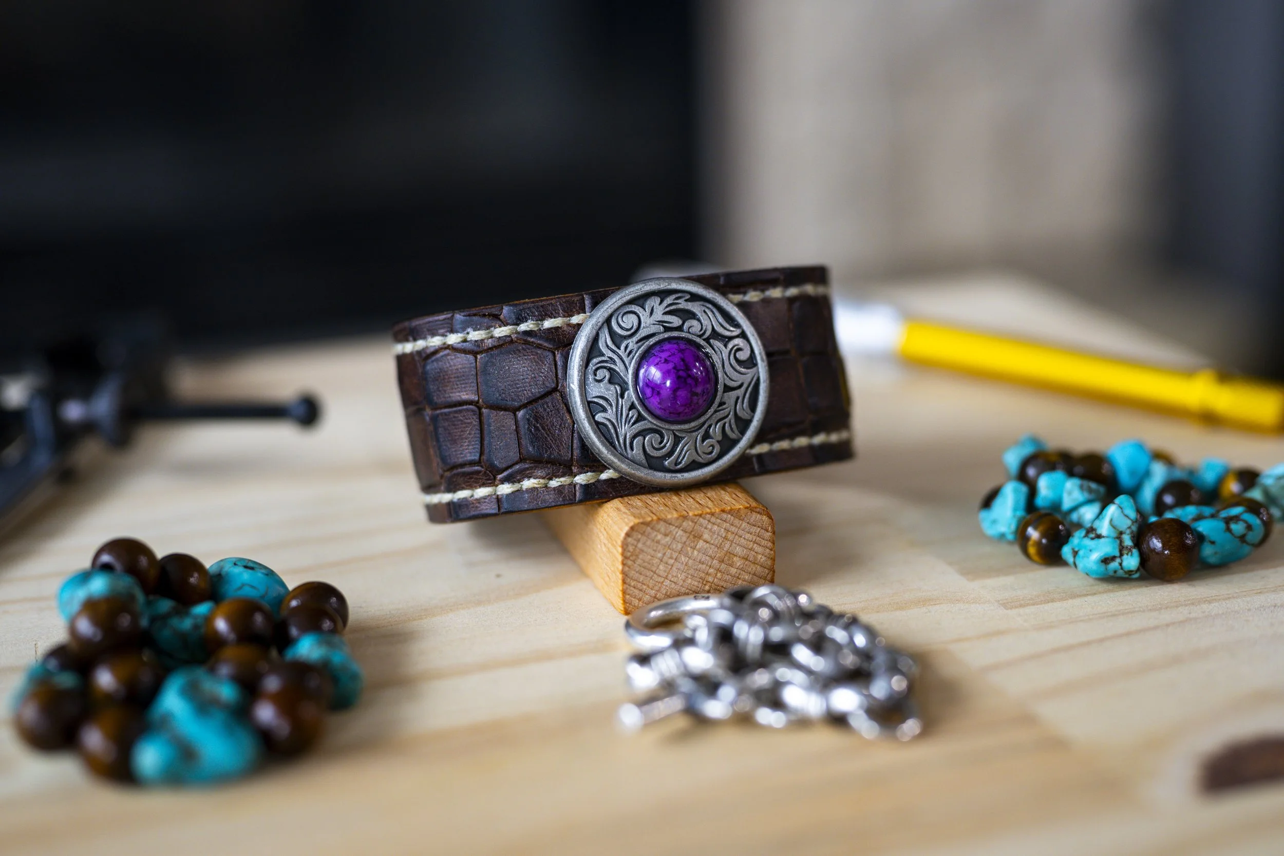 A brown leather bracelet with intricate silver detailing and a purple gemstone in the center, on a wooden stand, with turquoise and brown bead necklaces and a yellow pen in the background.