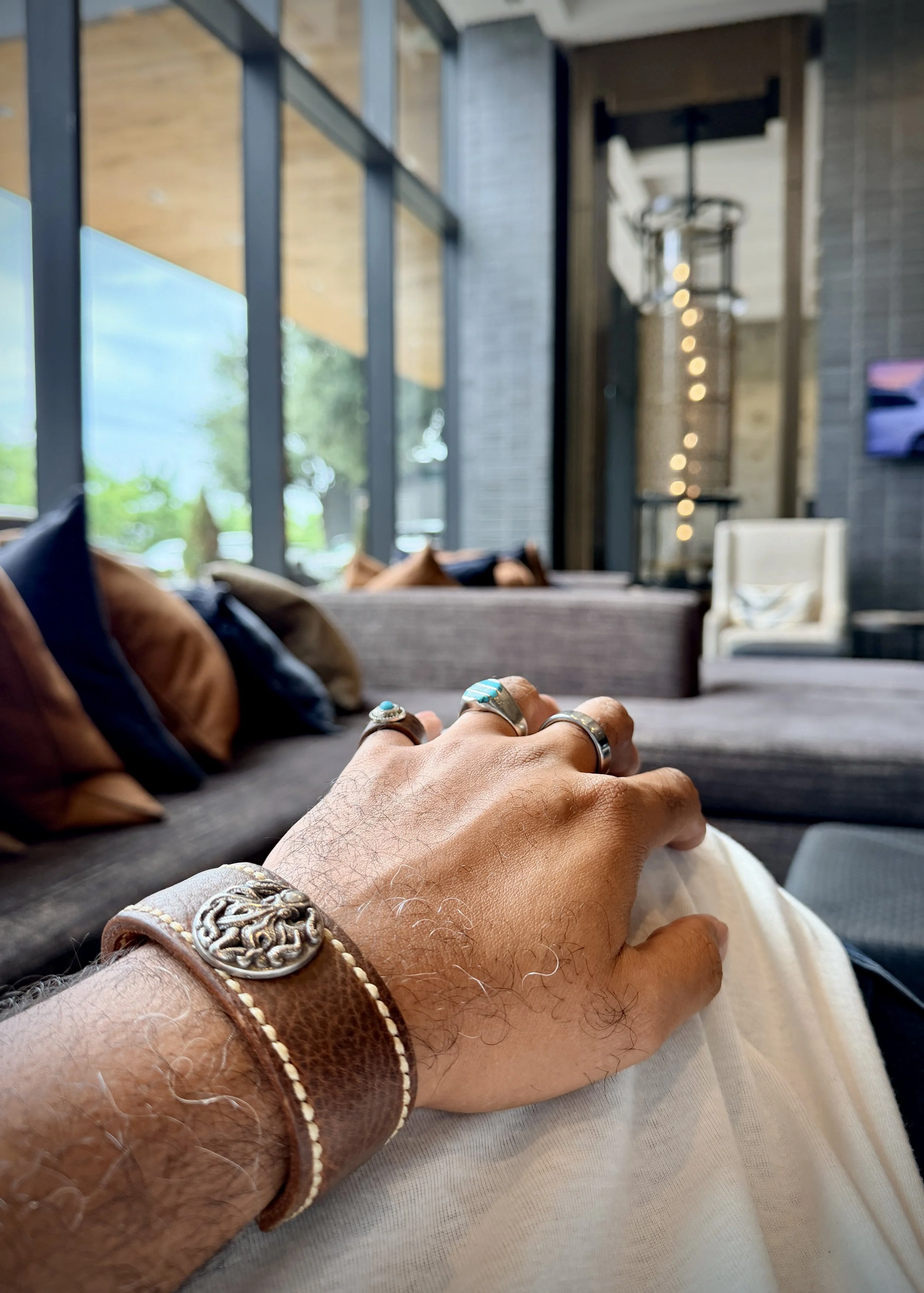 A hand wearing the Hilo leather cuff with an octopus concho and several turquoise rings rests on a knee inside a bright hotel lounge with large windows and seating in the background.