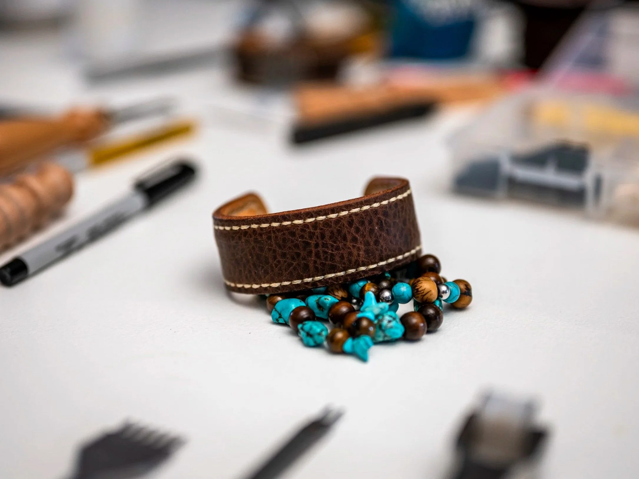 A handcrafted brown leather cuff with contrasting stitching sits above a cluster of turquoise, brown, and metallic beads, with leatherworking tools softly blurred across the workshop table behind it.