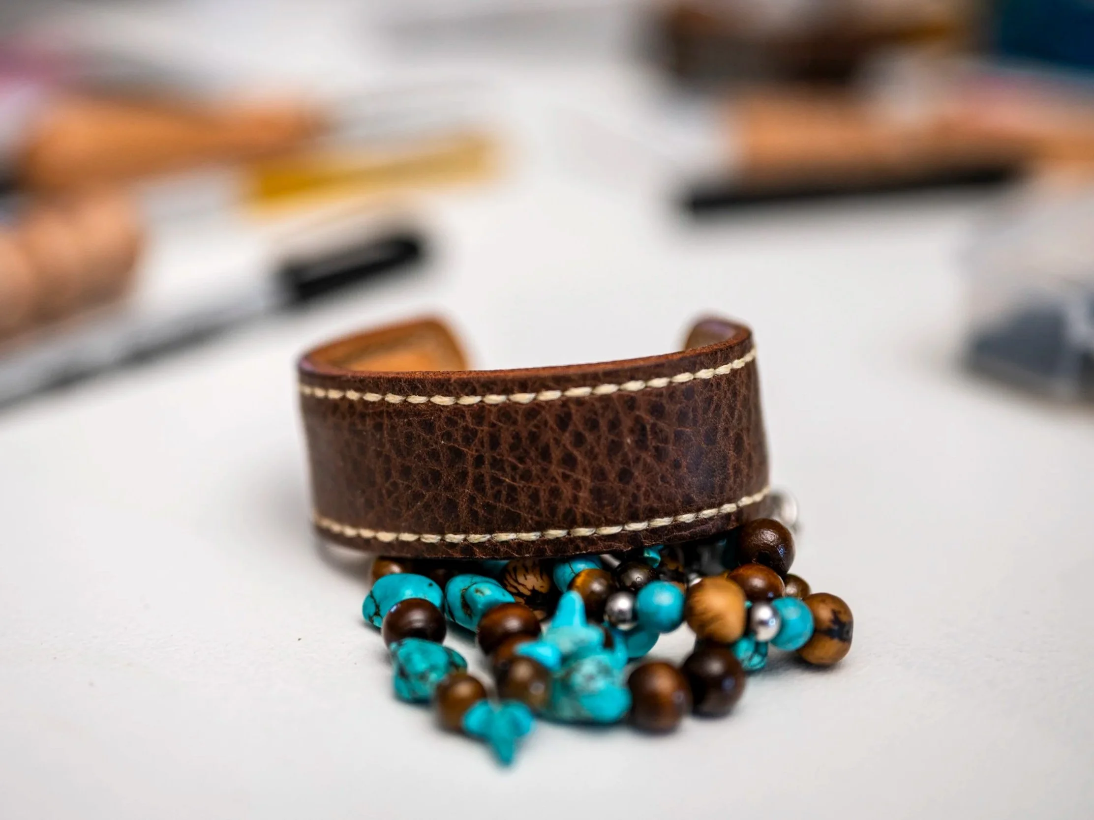 Close-up of a handcrafted brown leather cuff with contrasting stitching positioned above turquoise, brown, and metallic beads, with the workshop softly blurred in the background.