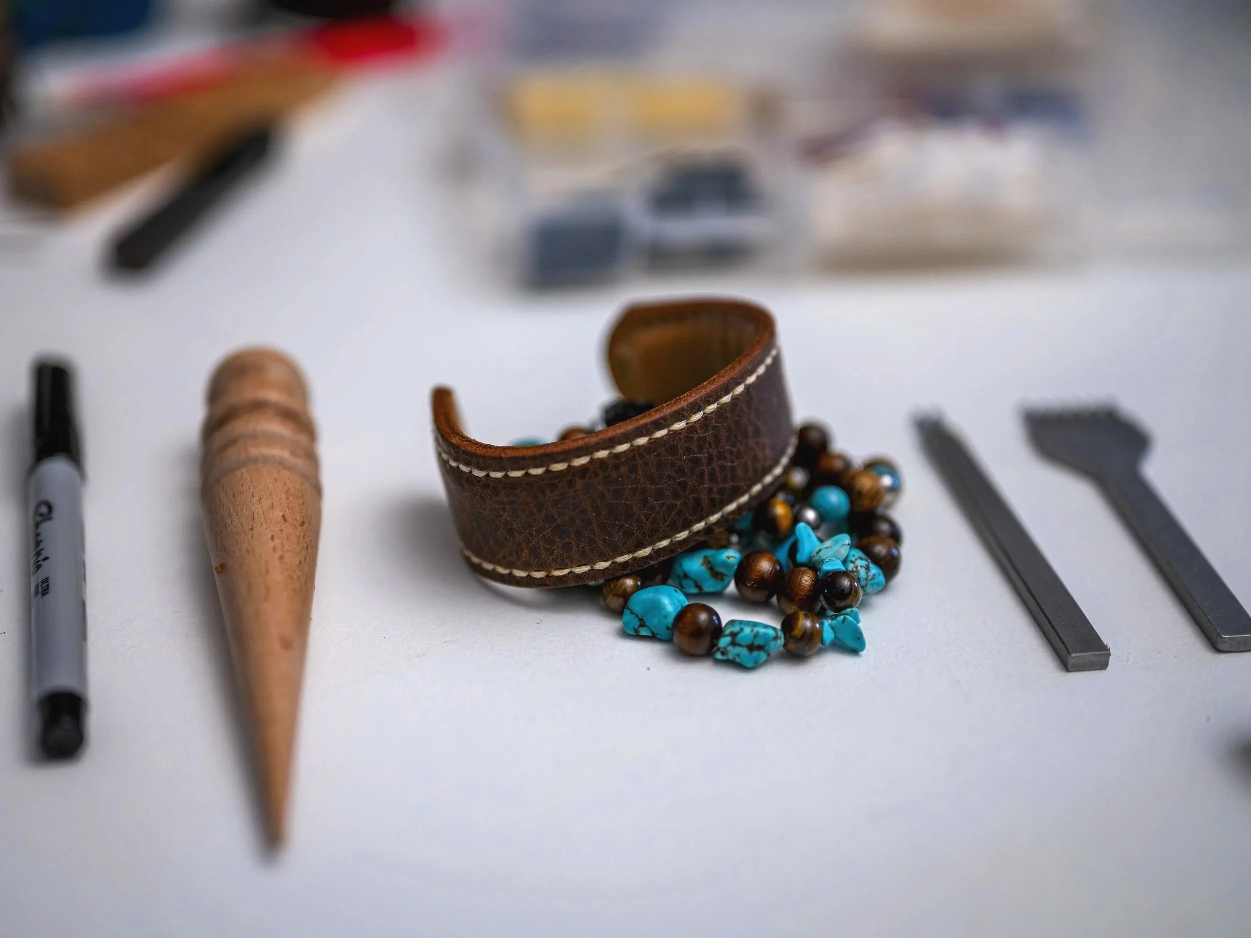 A handcrafted brown leather cuff with contrasting stitching sits among turquoise, brown, and metallic beads and leatherworking tools on a workshop table.