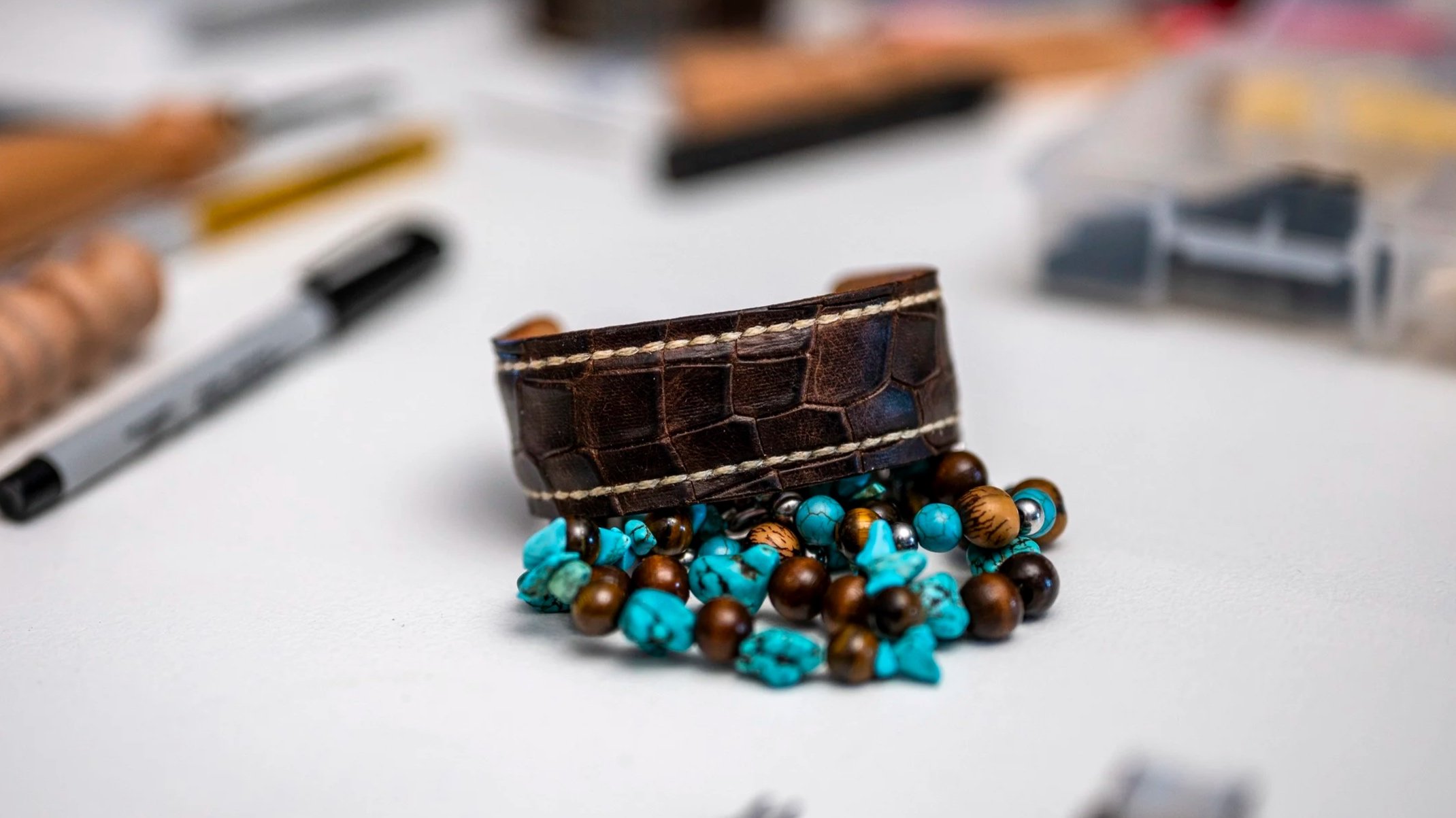 Close-up of a handcrafted brown leather cuff with contrasting stitching positioned above a cluster of turquoise, brown, and metallic beads, with leatherworking tools softly blurred in the background.