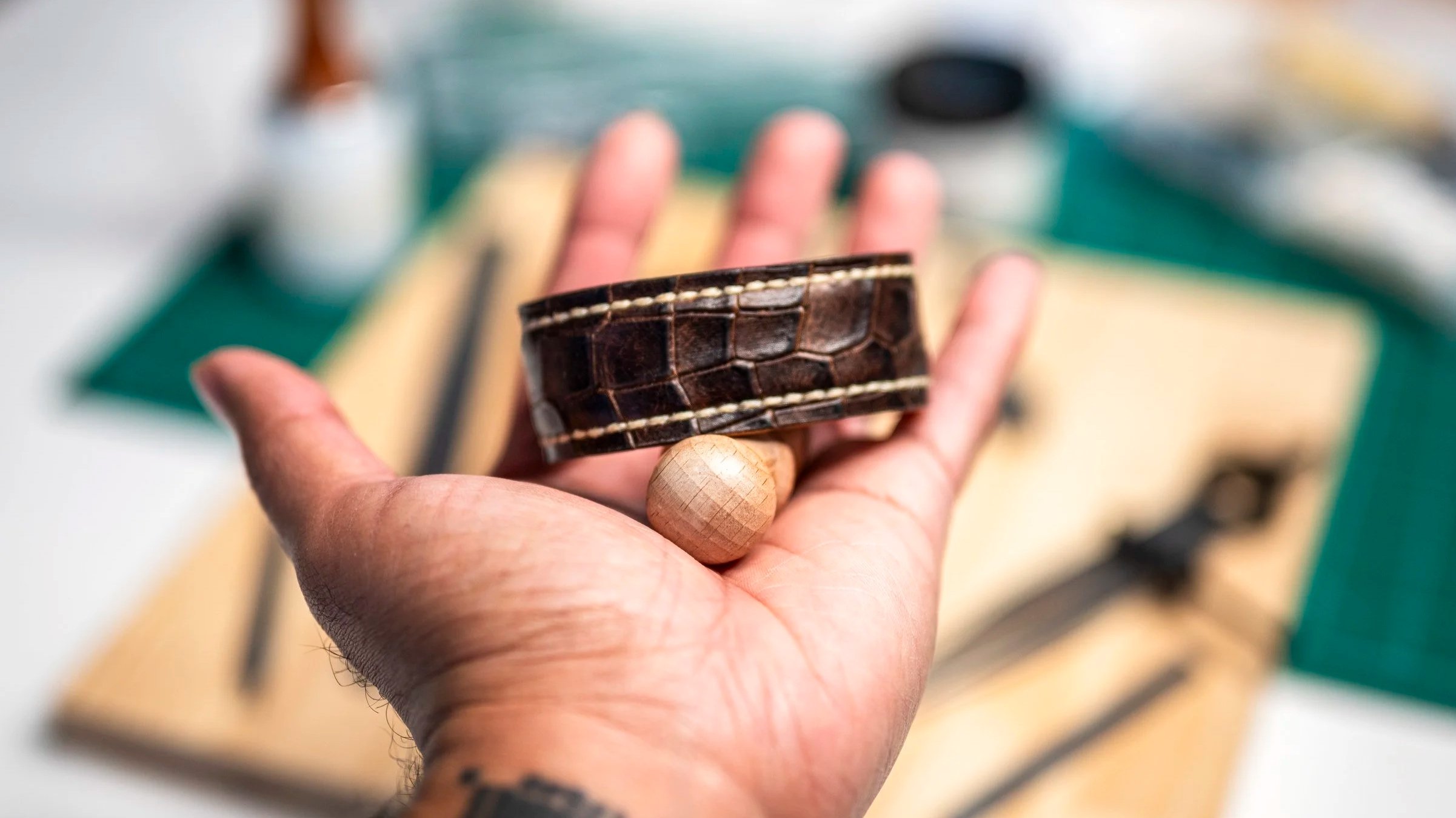 A Black hand holds a handcrafted brown leather cuff open to show its finished interior, contrasting stitching, and layered leather construction, with the workshop softly blurred behind it.