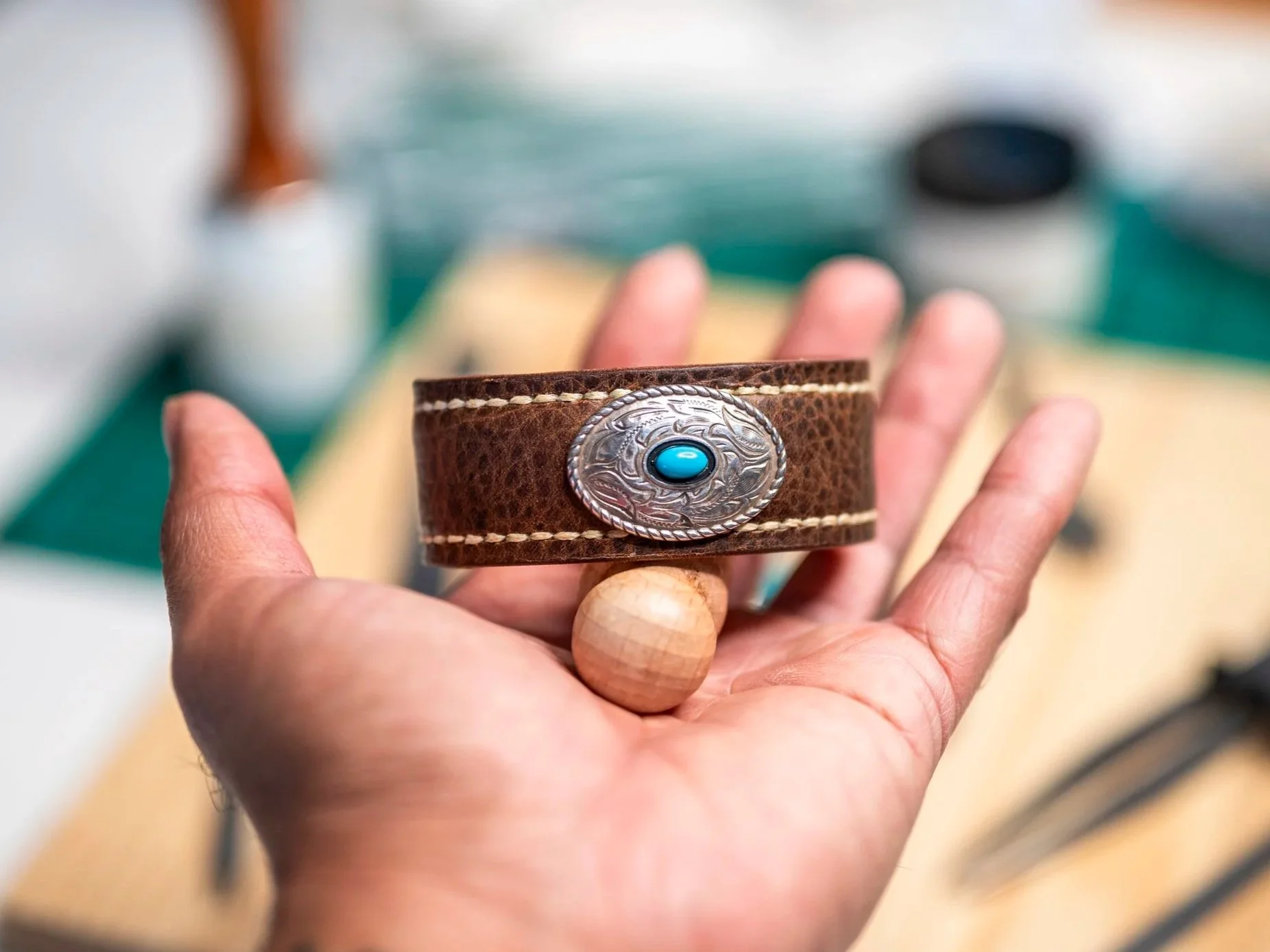 A Black hand holds the handcrafted Moana brown leather cuff, featuring white stitching and an oval silver-toned concho with a turquoise-colored center, against a softly blurred workshop background.