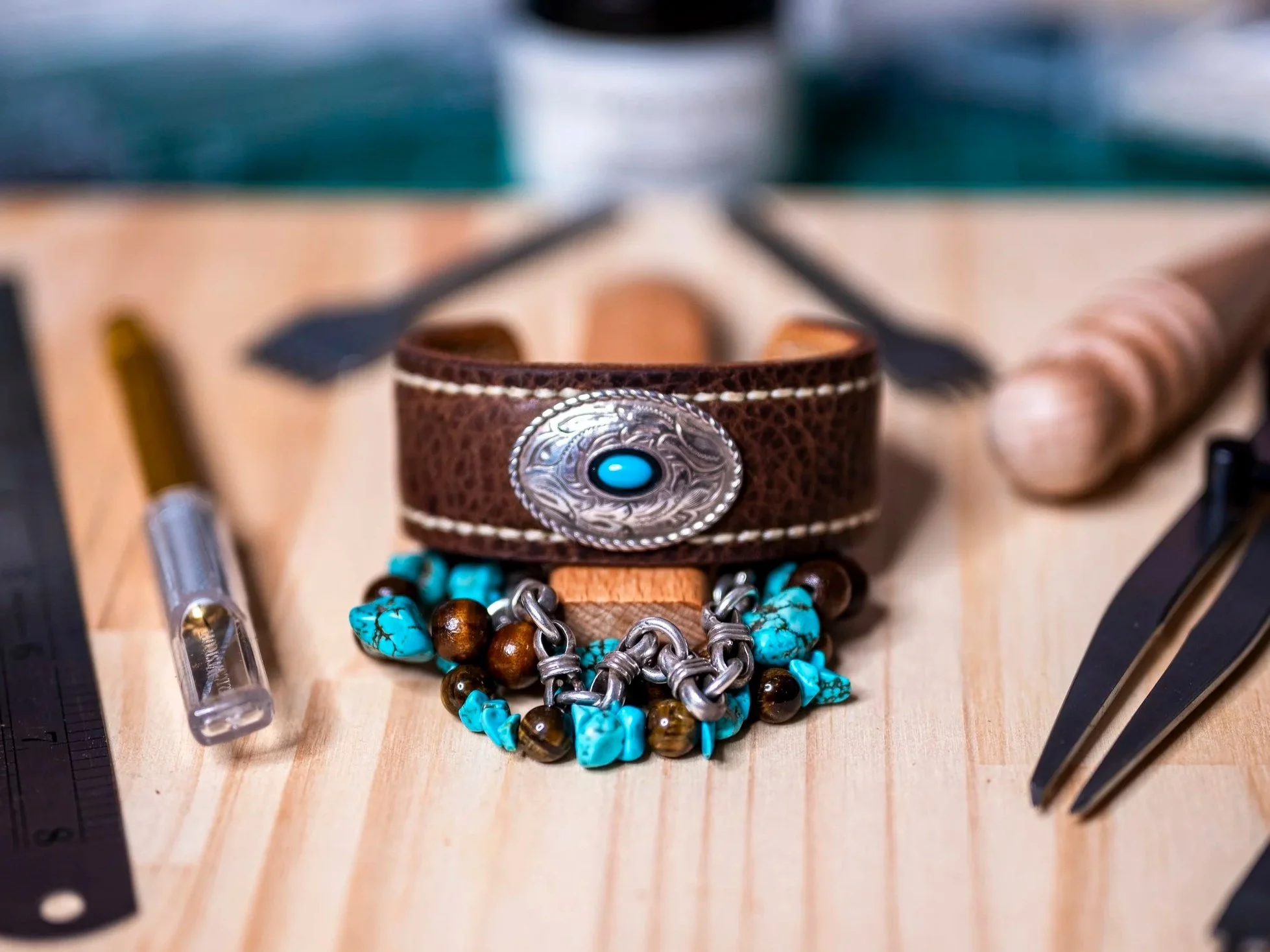Front view of the handcrafted Moana brown leather cuff, featuring an oval silver-toned concho with a turquoise-colored center, displayed with turquoise and brown beaded jewelry among leatherworking tools on a wooden workbench.