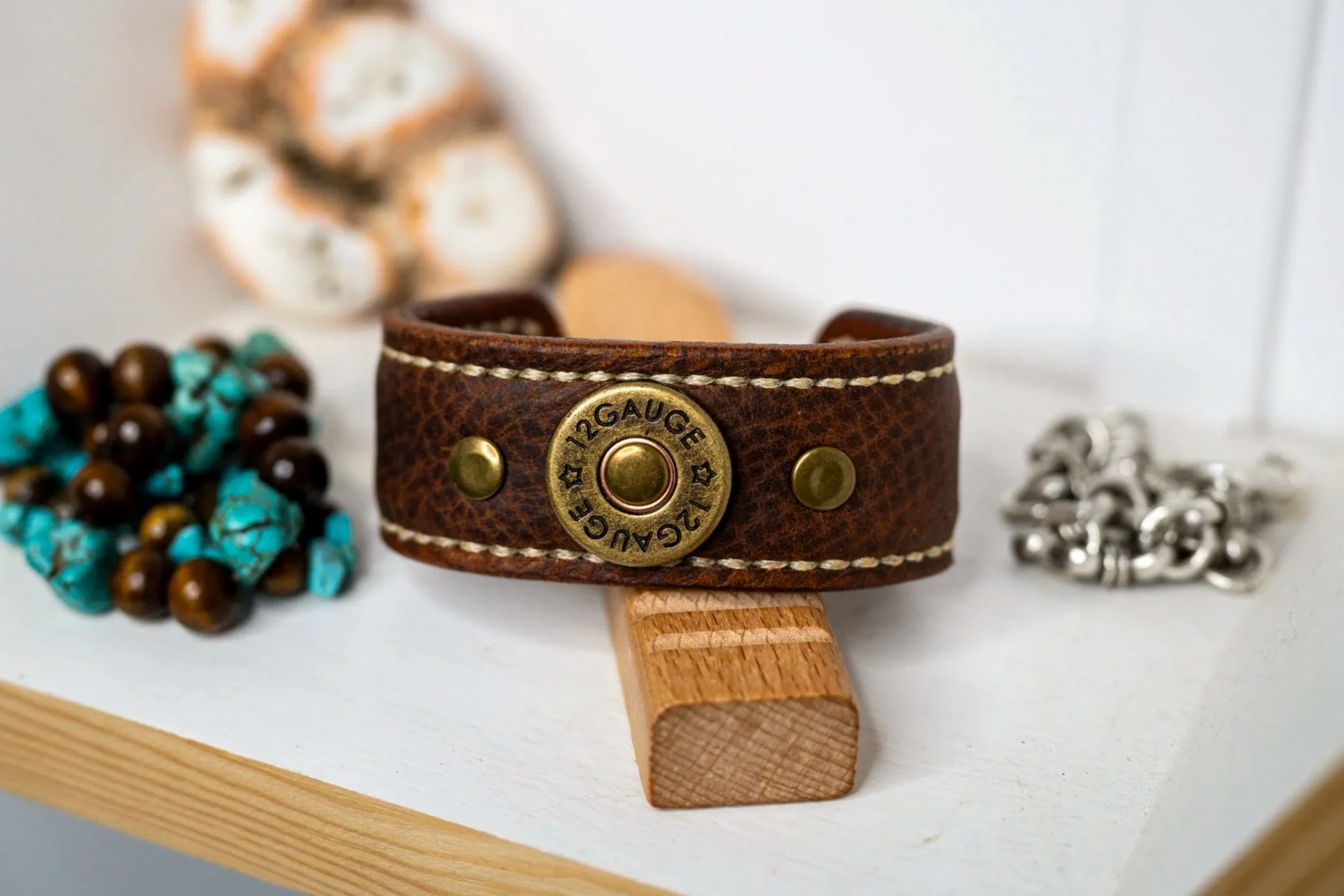 The Koa handcrafted brown leather cuff, featuring a round brass-toned concho and contrasting stitching, is displayed on a wooden stand with turquoise-colored beads and silver-toned materials arranged nearby.