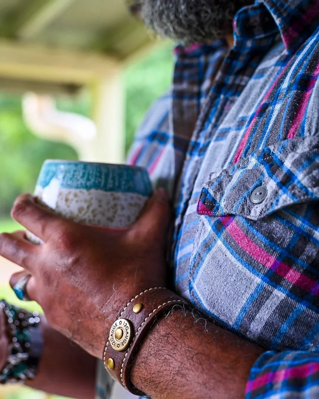 A Black man in a blue and red plaid shirt holds a ceramic cup outdoors while wearing the handcrafted Koa brown leather cuff, with soft greenery in the background.