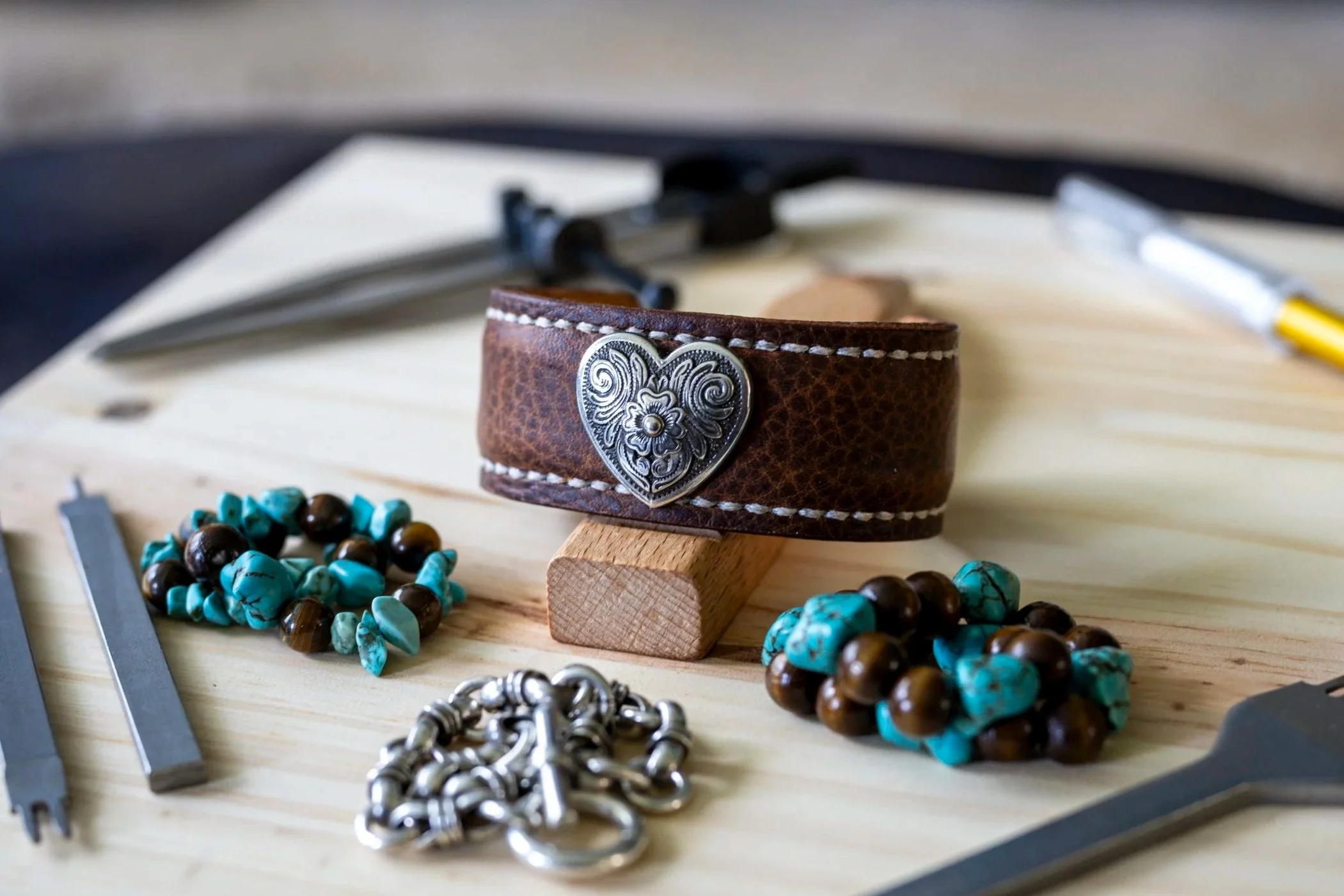 The handcrafted Waimea brown leather cuff displayed on a wooden stand, featuring white stitching and an ornate silver-toned heart concho, surrounded by turquoise, brown, and silver-toned jewelry on a workshop surface.