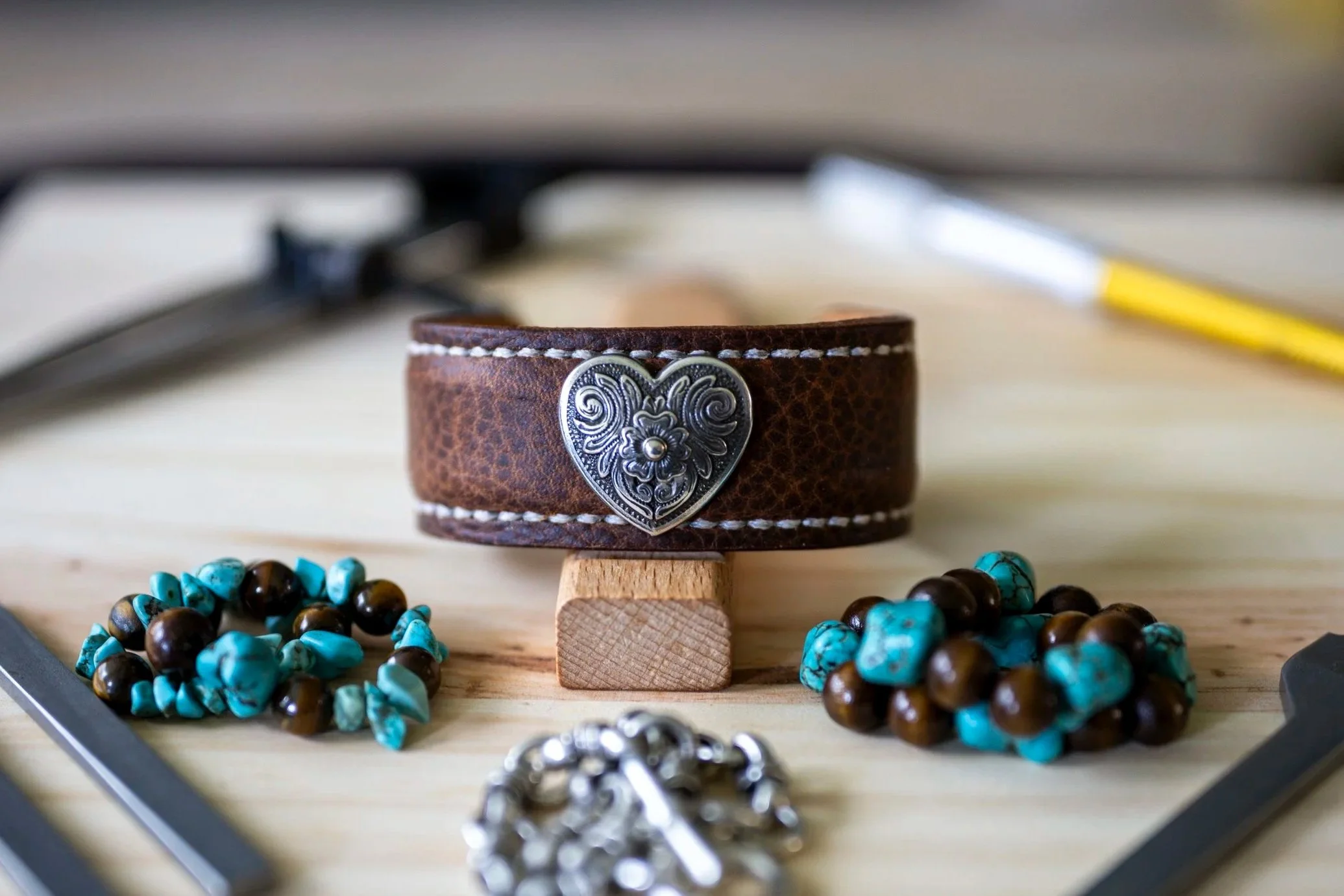 A close-up of a brown leather bracelet with a silver decorative heart-shaped emblem, resting on small wooden blocks, surrounded by turquoise and brown beaded necklaces on a wooden surface.