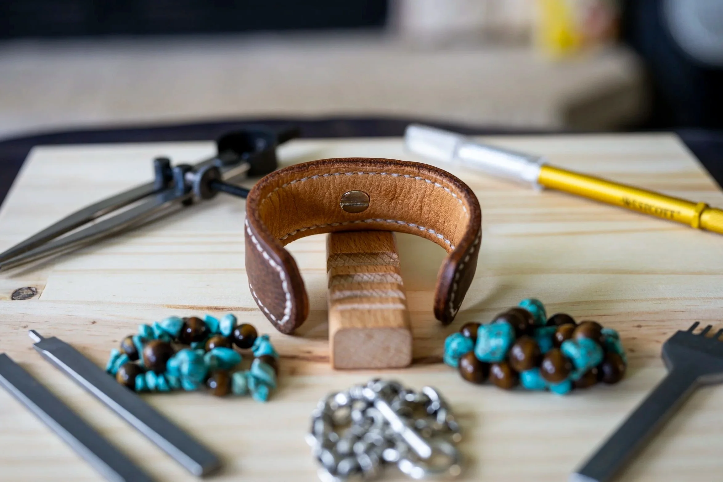 A wooden work surface with a leather bracelet, a metal compass, a yellow pen, and a beaded bracelet.