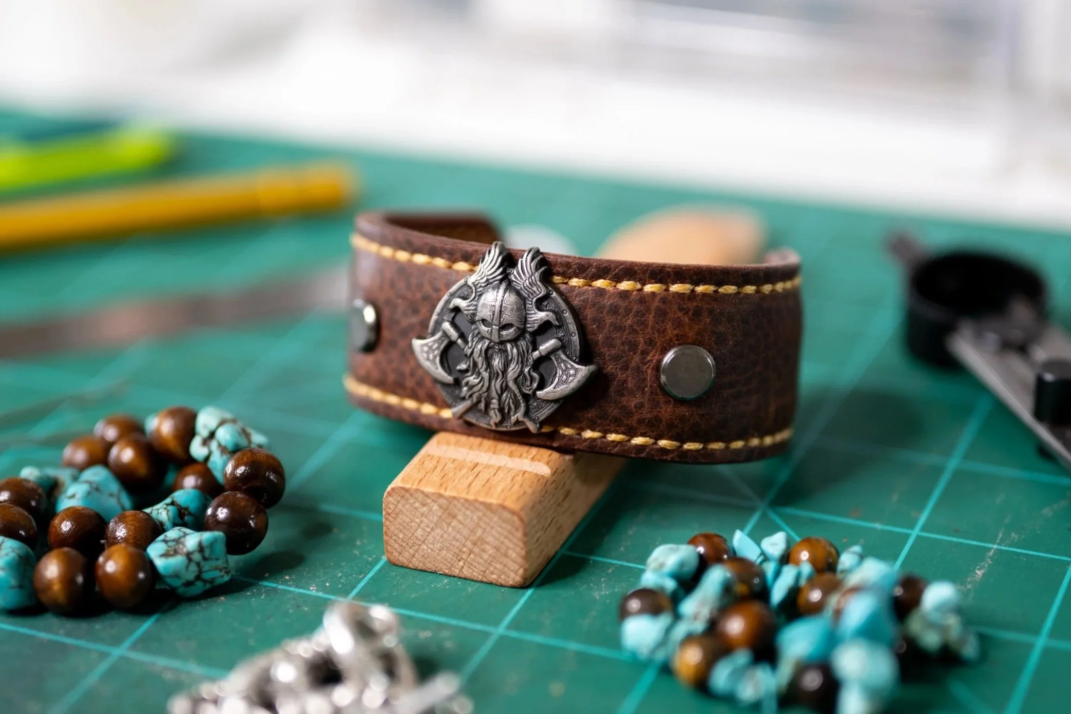 A close-up of a brown leather bracelet with a metal Viking dragon head medal, resting on a wooden block on a green cutting mat, with turquoise and brown beaded jewelry scattered around.