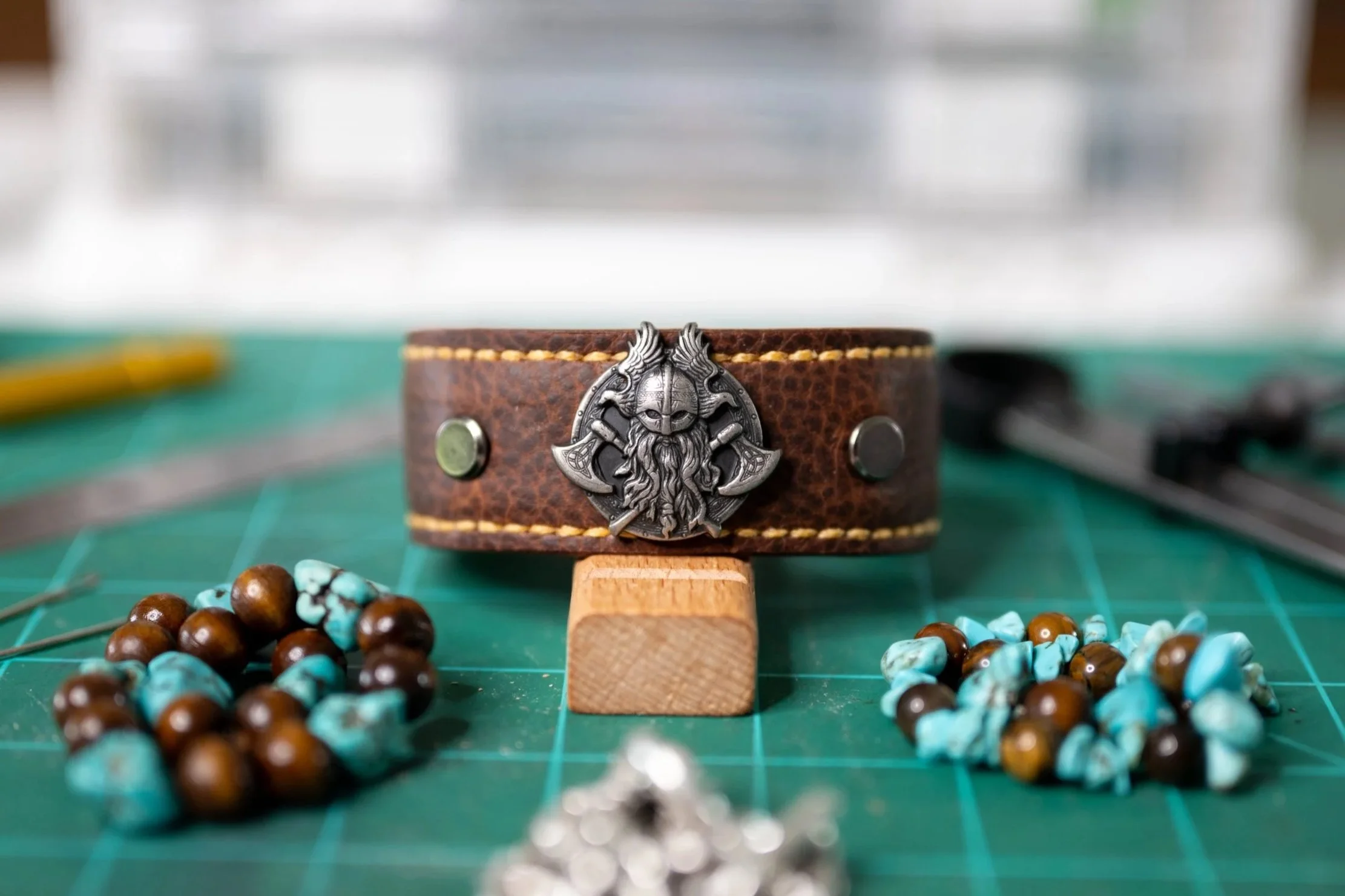 Close-up of a brown leather bracelet with a silver Viking helmet emblem on a green cutting mat. Beaded jewelry made of turquoise and brown beads is scattered around it, with some beads out of focus in the foreground.