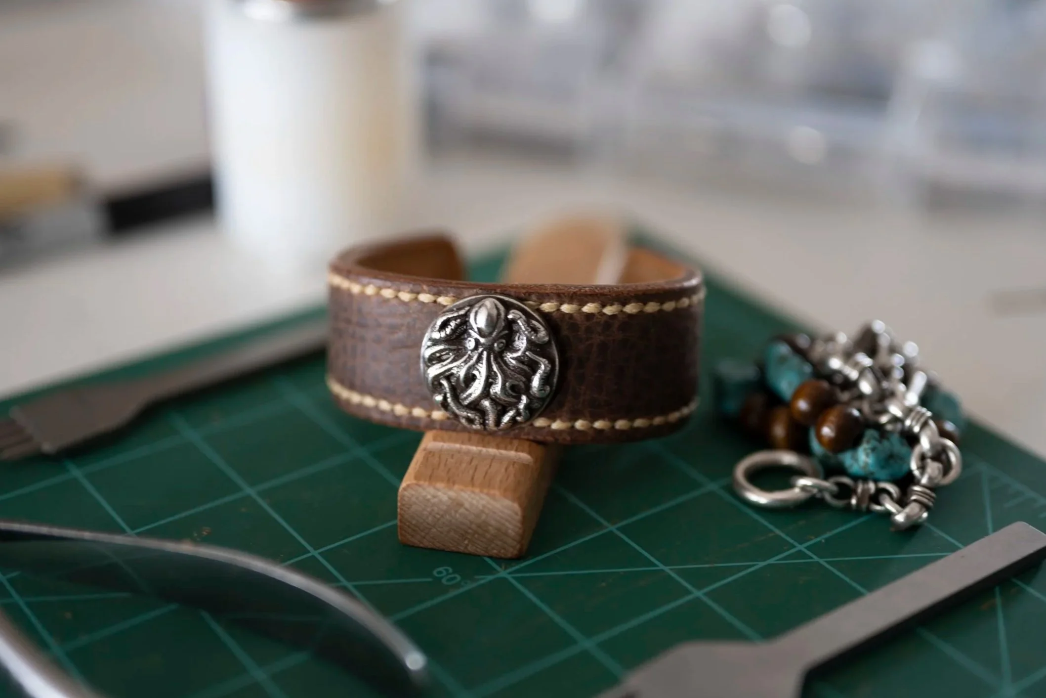 A brown leather bracelet with a metal decorative emblem, placed on a small wooden modeling stand on a green cutting mat. There is also a silver and turquoise beaded bracelet nearby, partially visible.