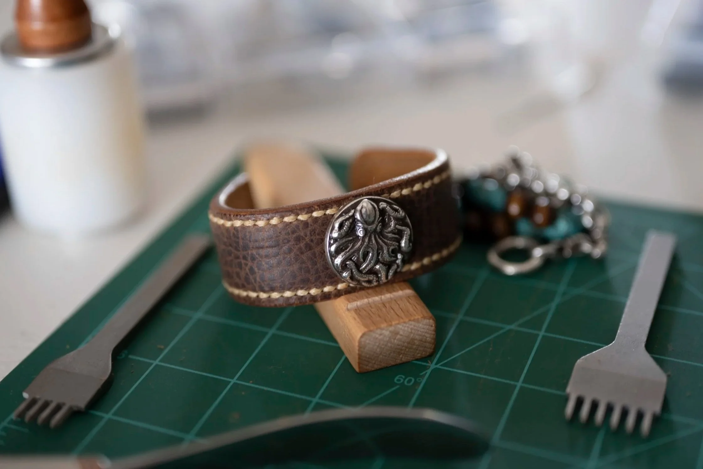 Close-up of a brown leather bracelet with a decorative metallic emblem, resting on a green cutting mat alongside a cutting tool with a black handle and a pair of pliers, in a workshop or crafting space.