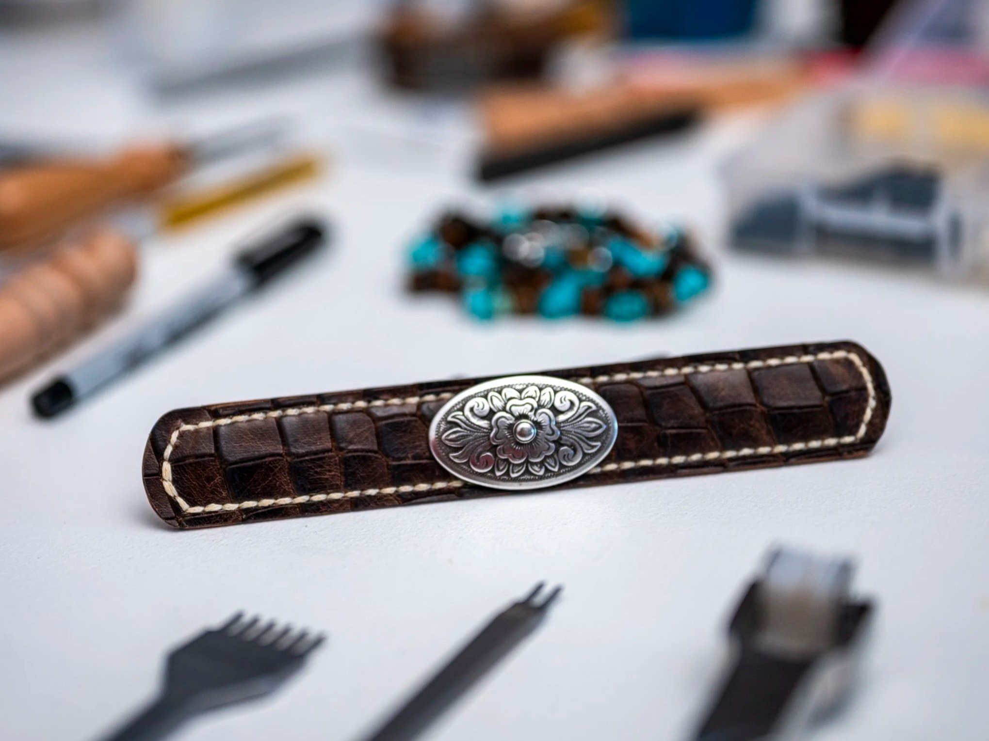 A close-up view of a brown leather wallet with decorative silver embellishment, with blurred background including various small objects and a pile of pills or beads on a white surface.