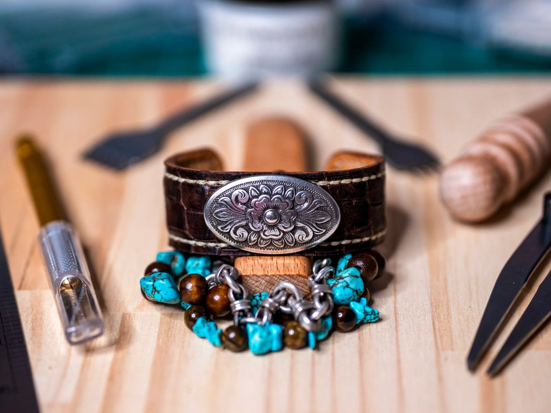 Close-up of a leather bracelet with a silver floral emblem, turquoise and brown beaded necklaces, and other jewelry items on a wooden surface.