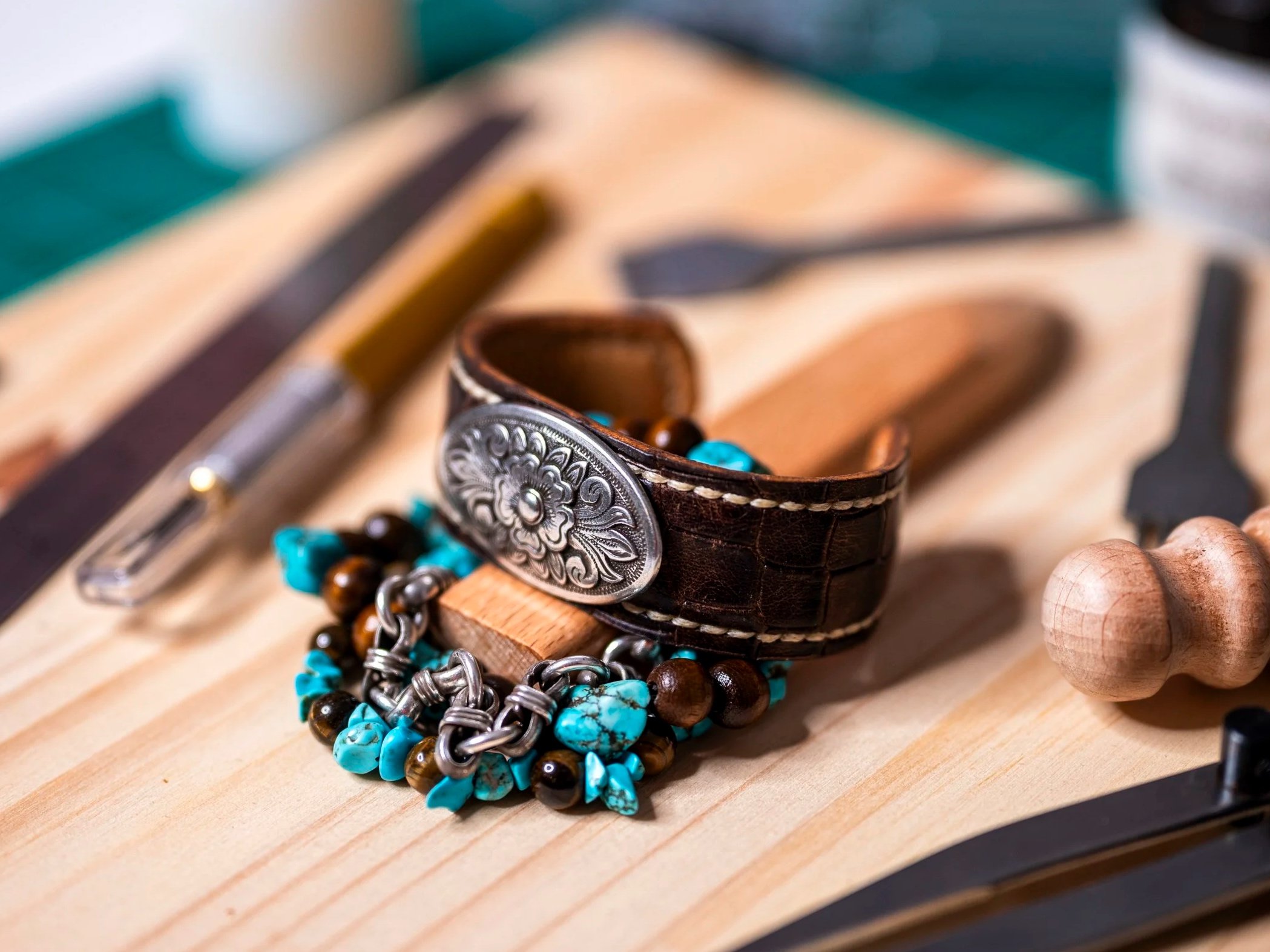 Close-up of a brown leather bracelet with engraved silver medallion, turquoise and brown beaded bracelets, and a silver chain bracelet on a wooden surface.