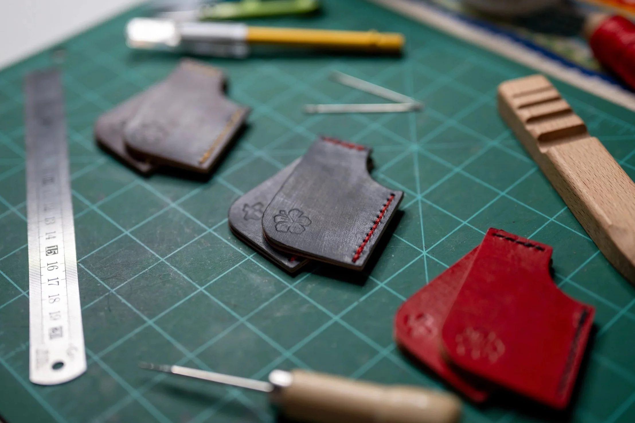 A green cutting mat with various craft supplies, including small fabric pieces with embroidered designs, a metal ruler, yellow paintbrush, two metal clips, a wooden block, and a partially visible red fabric piece on the right.