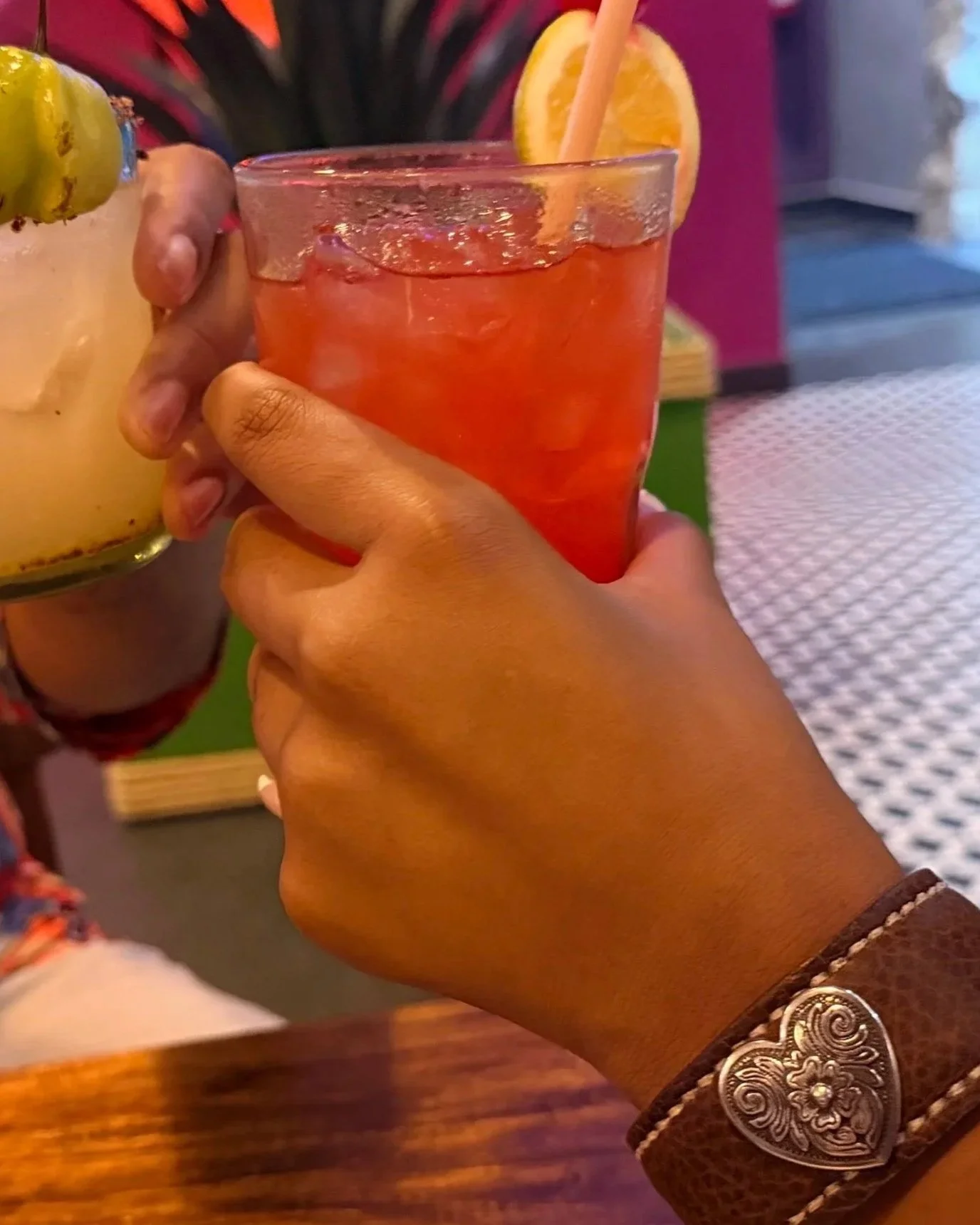 A wrist wearing the Waimea leather cuff rests near a table as two people hold colorful drinks together during an evening out, creating a casual shared moment.