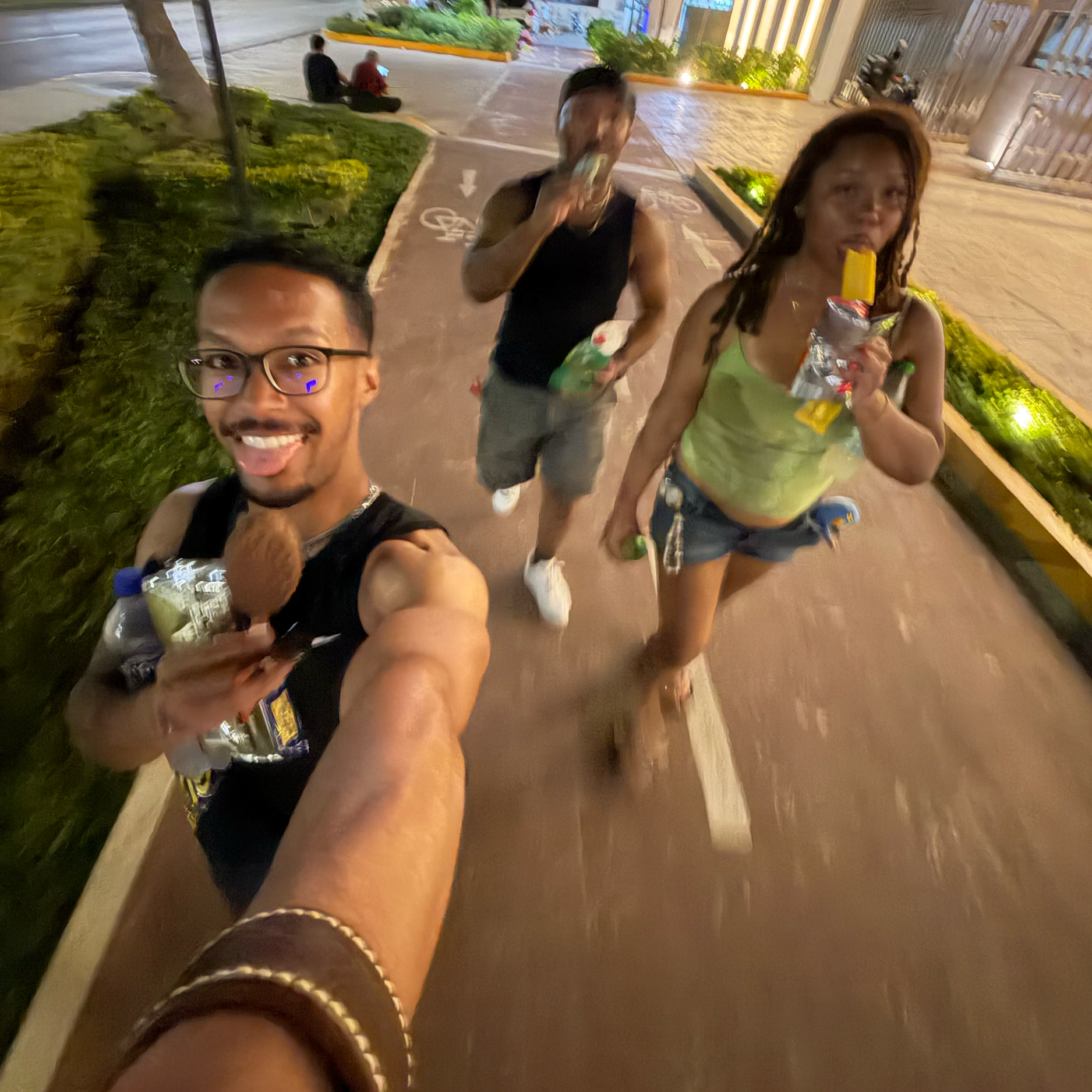 A smiling Black man wearing the Makahā leather cuff takes a wide-angle selfie with two friends of color during an evening out in Cancún, Mexico, along a brightly lit outdoor walkway surrounded by tropical greenery.