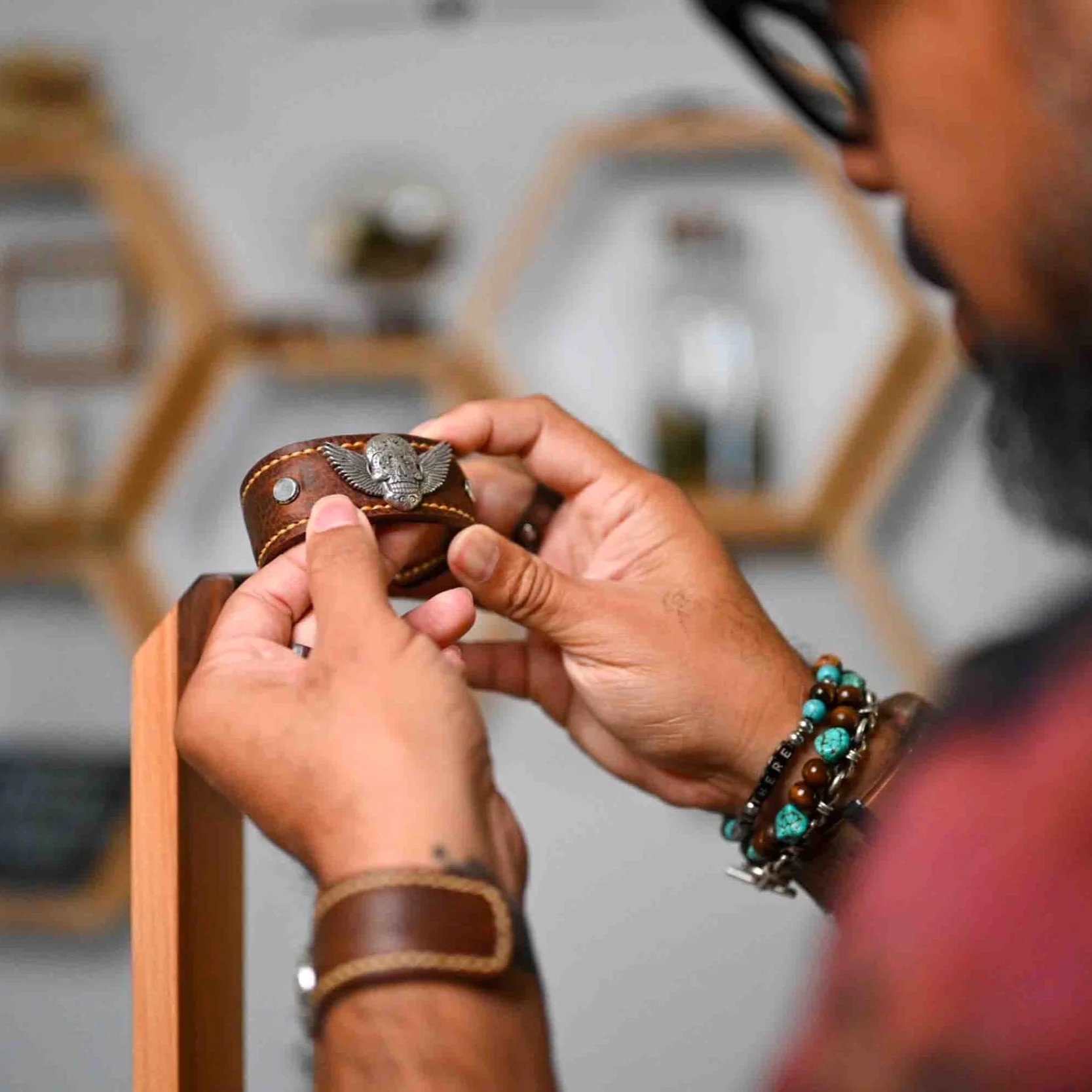 A person holding a leather bracelet with a silver winged skull embellishment, wearing multiple beaded bracelets on their wrist, with blurred furniture in the background.
