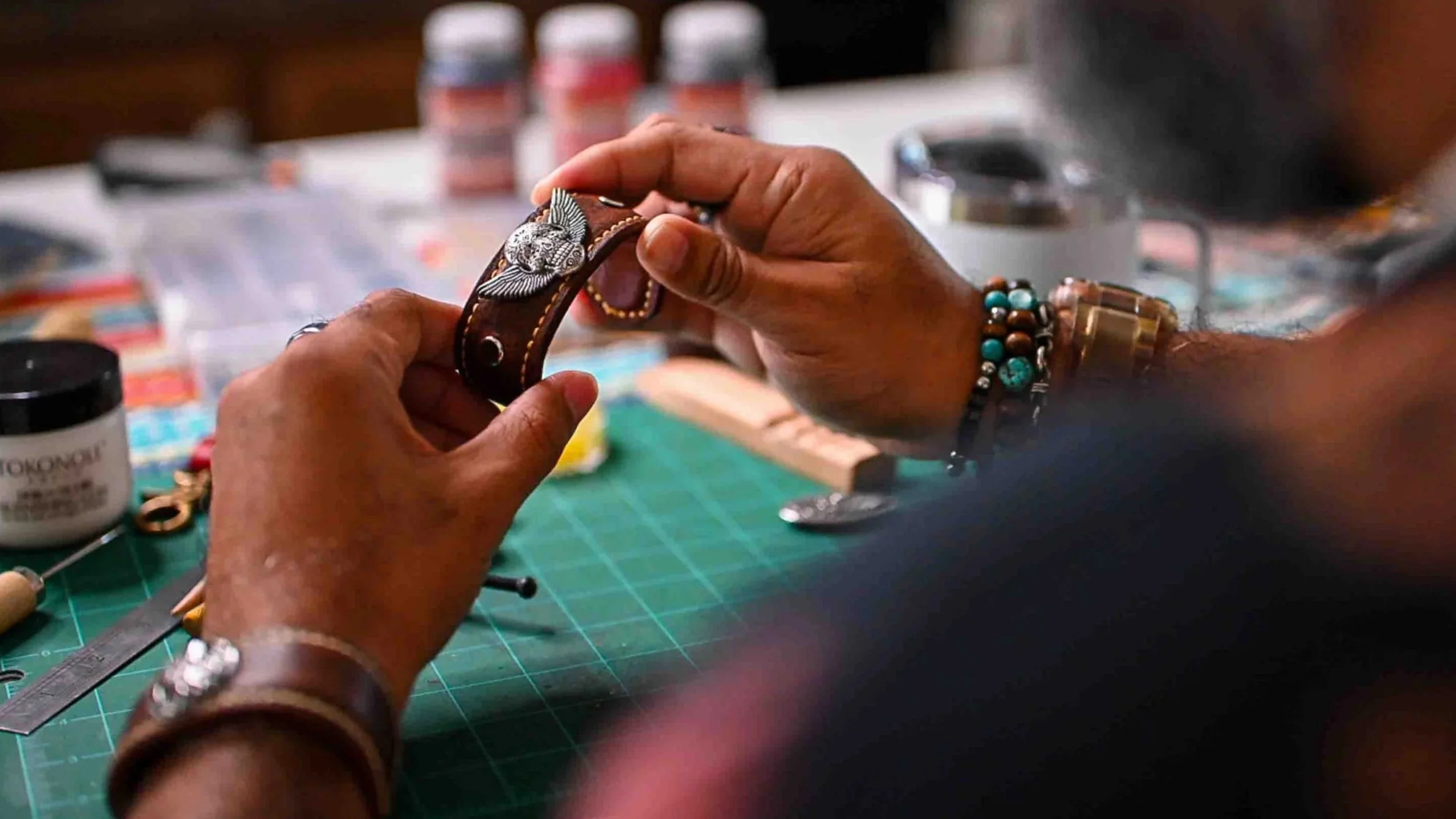 Person holding a leather bracelet with a silver fish embellishment, surrounded by jewelry-making supplies on a work table.