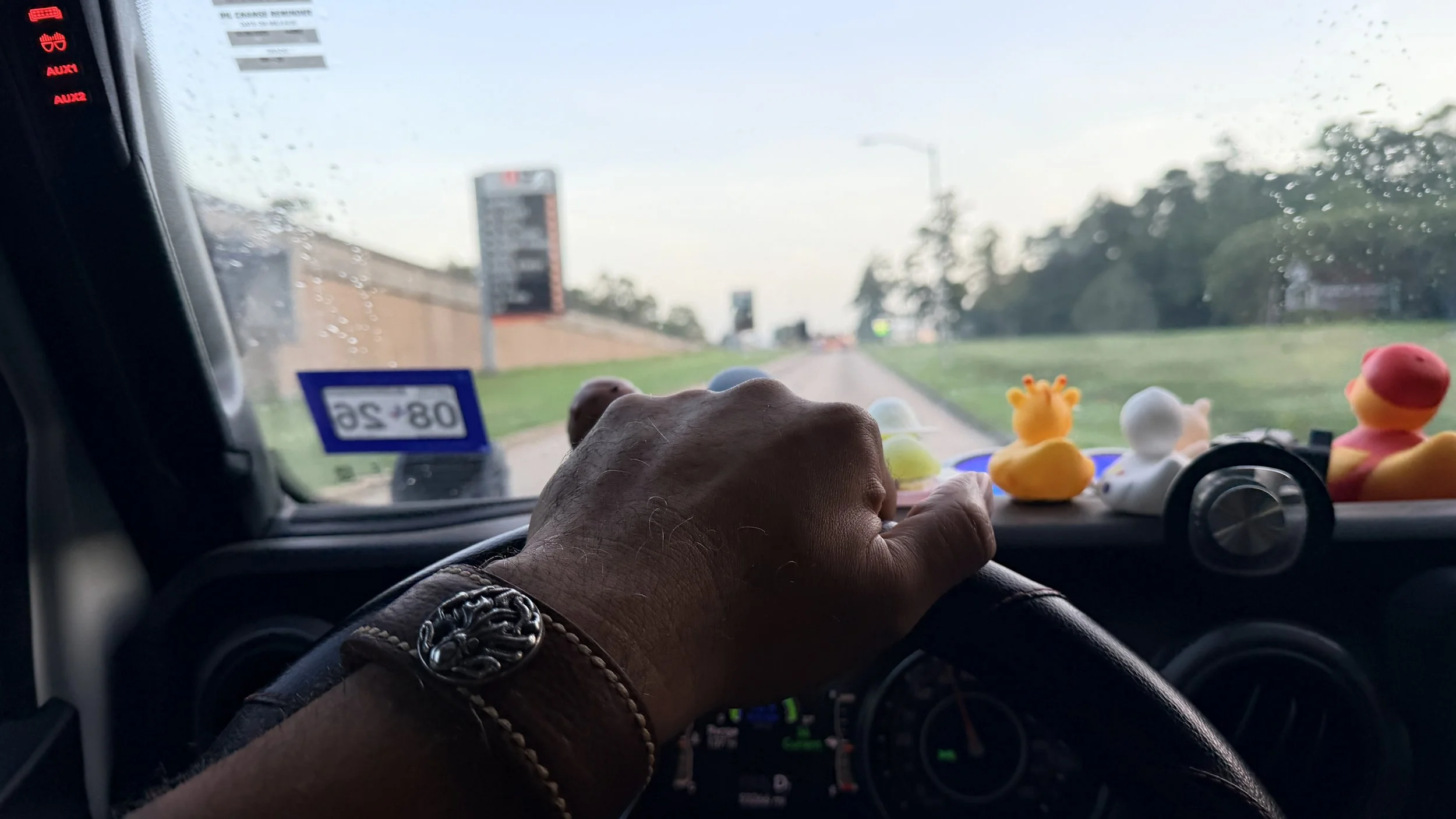 A Black man’s hand wearing the Hilo handcrafted brown leather cuff rests on the steering wheel while driving on a rain-dampened road, with colorful rubber ducks lined across the dashboard.