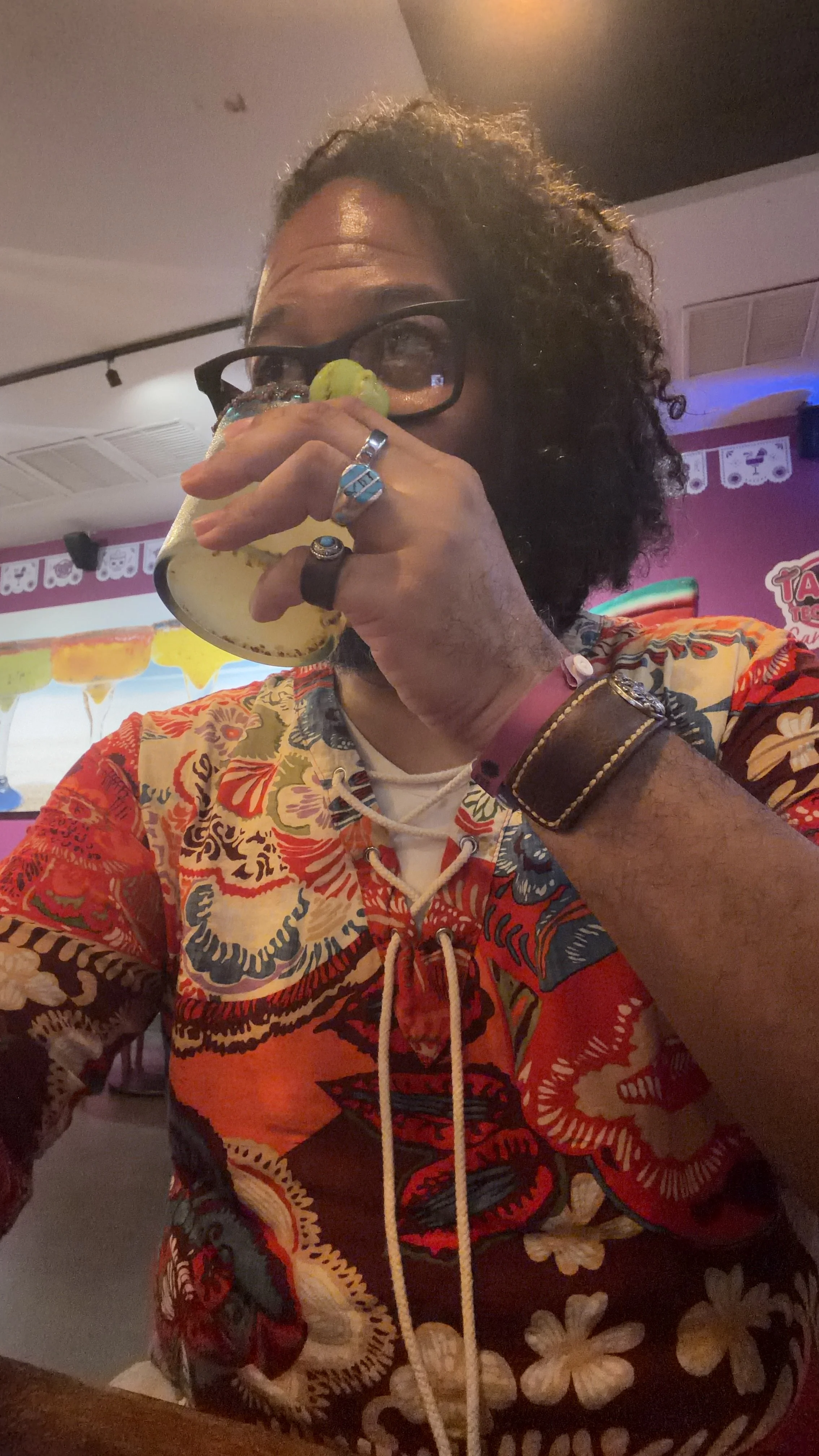 A Black man with curly micro locs and glasses, wearing a colorful patterned shirt, takes a sip from a drink while enjoying a lively indoor gathering.