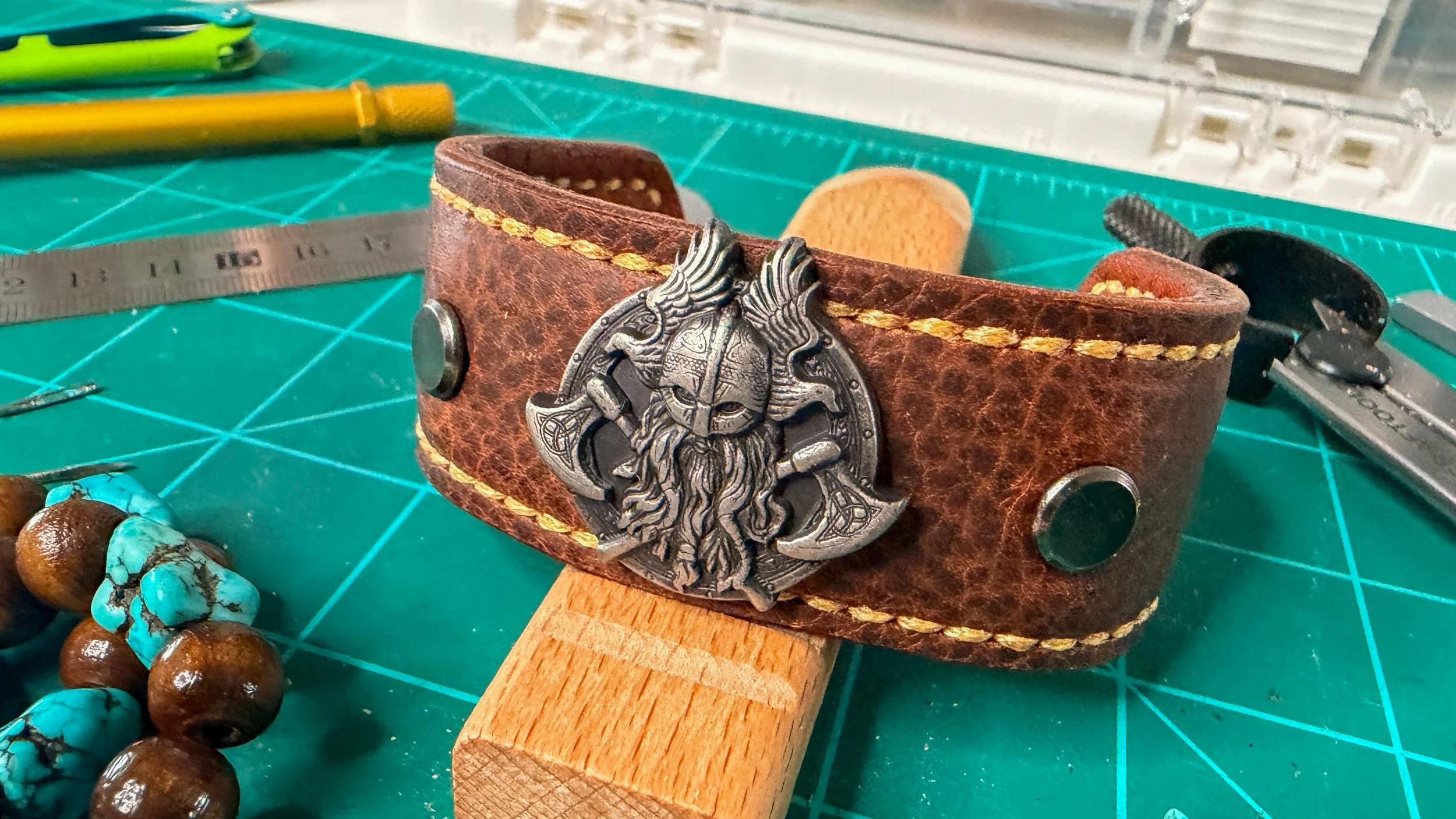 A leather strap bracelet with a Viking warrior head metal medallion, surrounded by jewelry-making tools and beads on a green cutting mat.