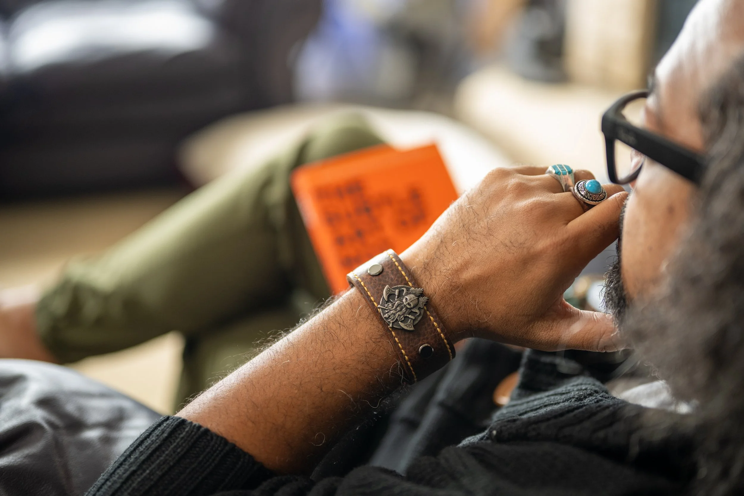 A person with glasses, wearing rings and bracelets, is holding a phone to their ear. They have a beard and are wearing a dark sweater.