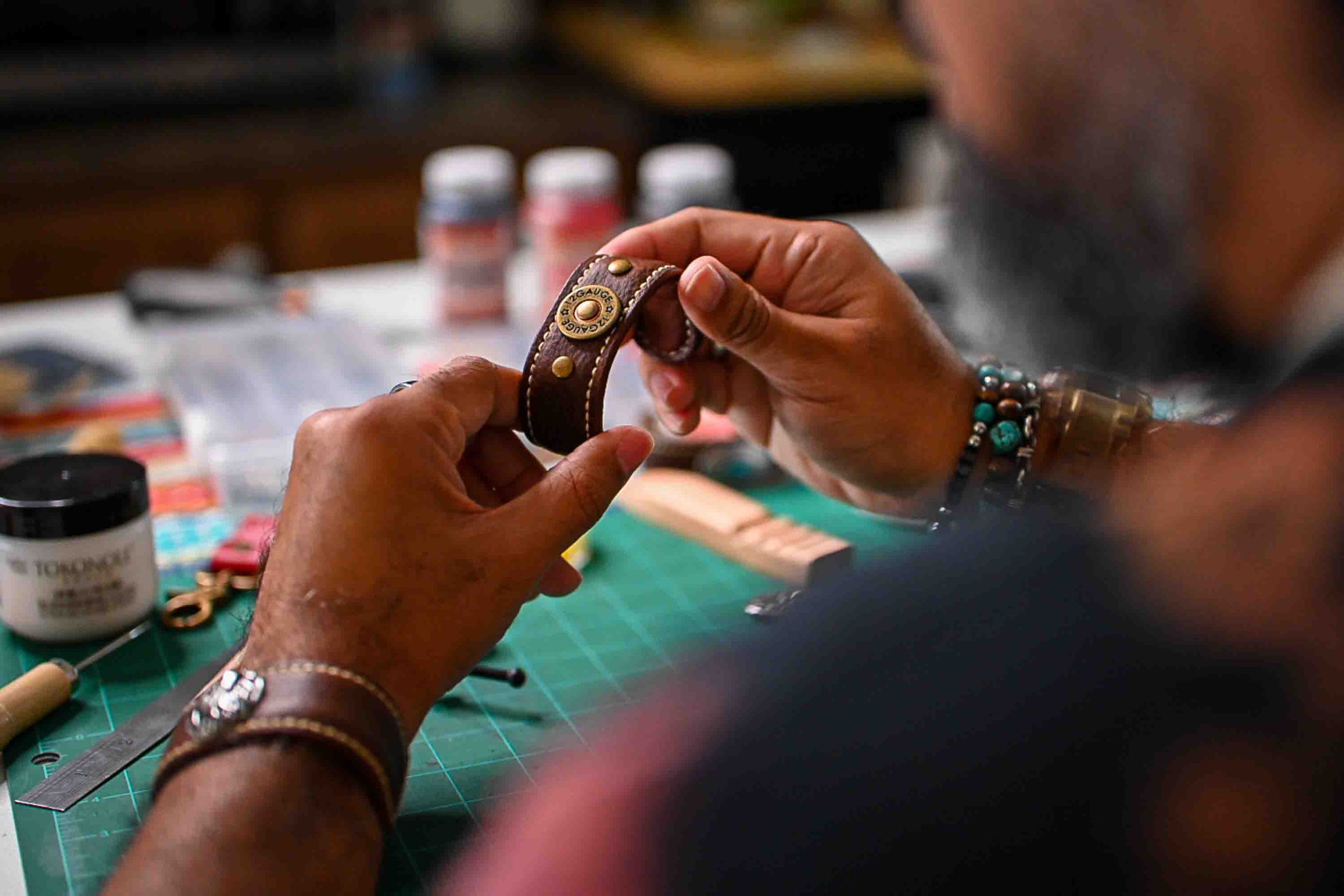 A Black leather artisan examines the handcrafted Koa cuff at his workshop table, surrounded by leatherworking tools and supplies on a green cutting mat.