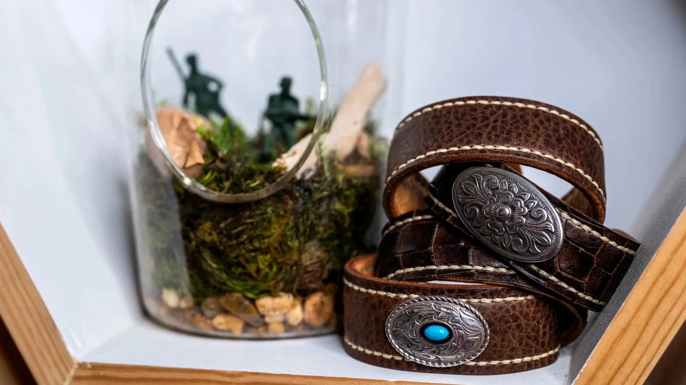 The Makahā, Kapahulu, and Moana handcrafted brown leather cuffs are stacked beside a glass terrarium filled with moss, small plants, and miniature figures, with Moana’s turquoise-accented concho visible in front.
