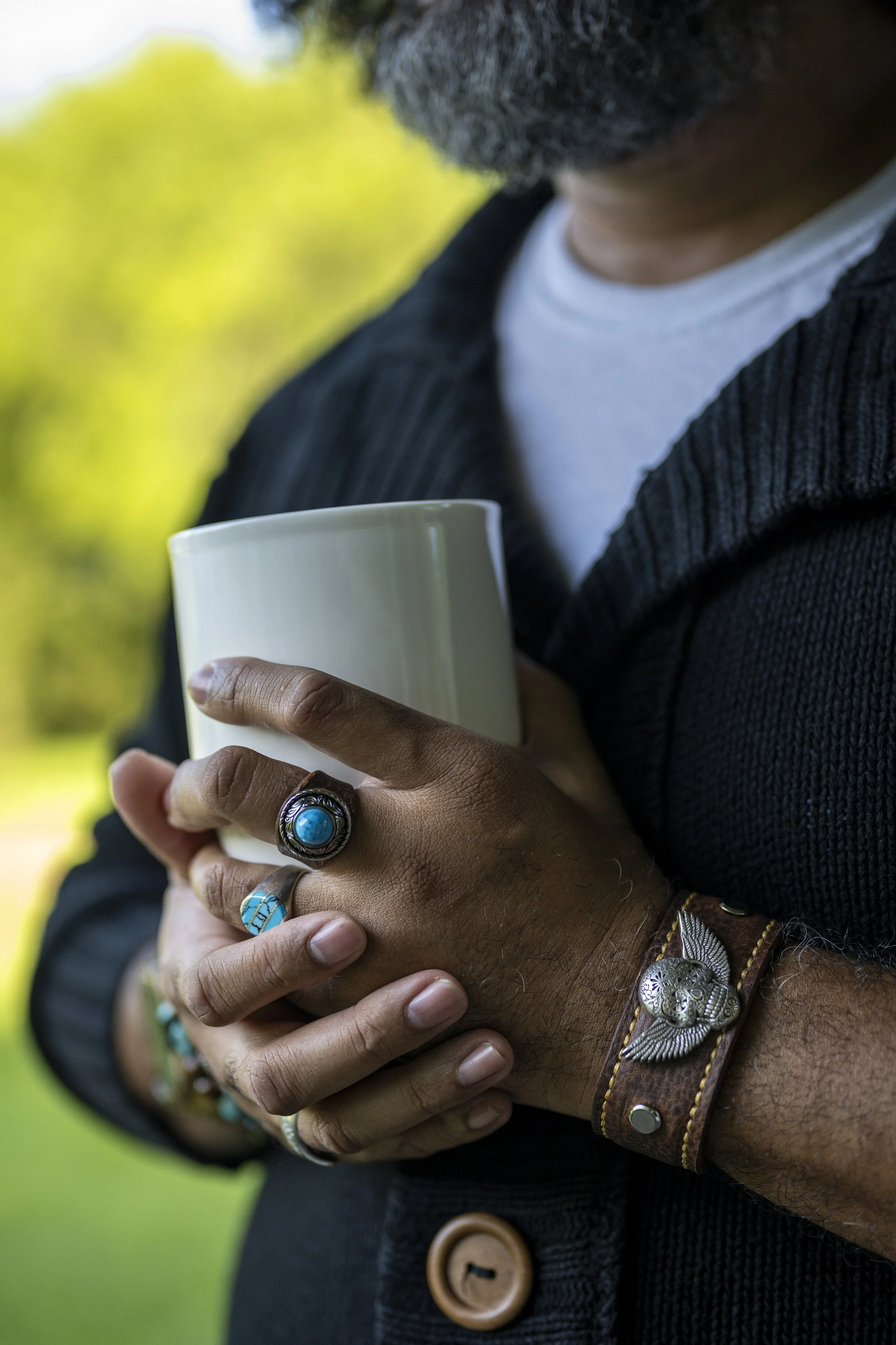 Close-up of a person holding a white mug, wearing a black corduroy jacket over a white shirt, with visible jewelry including a turquoise ring, a leather bracelet with metallic embellishments, and a silver feather-shaped bracelet, in an outdoor settin