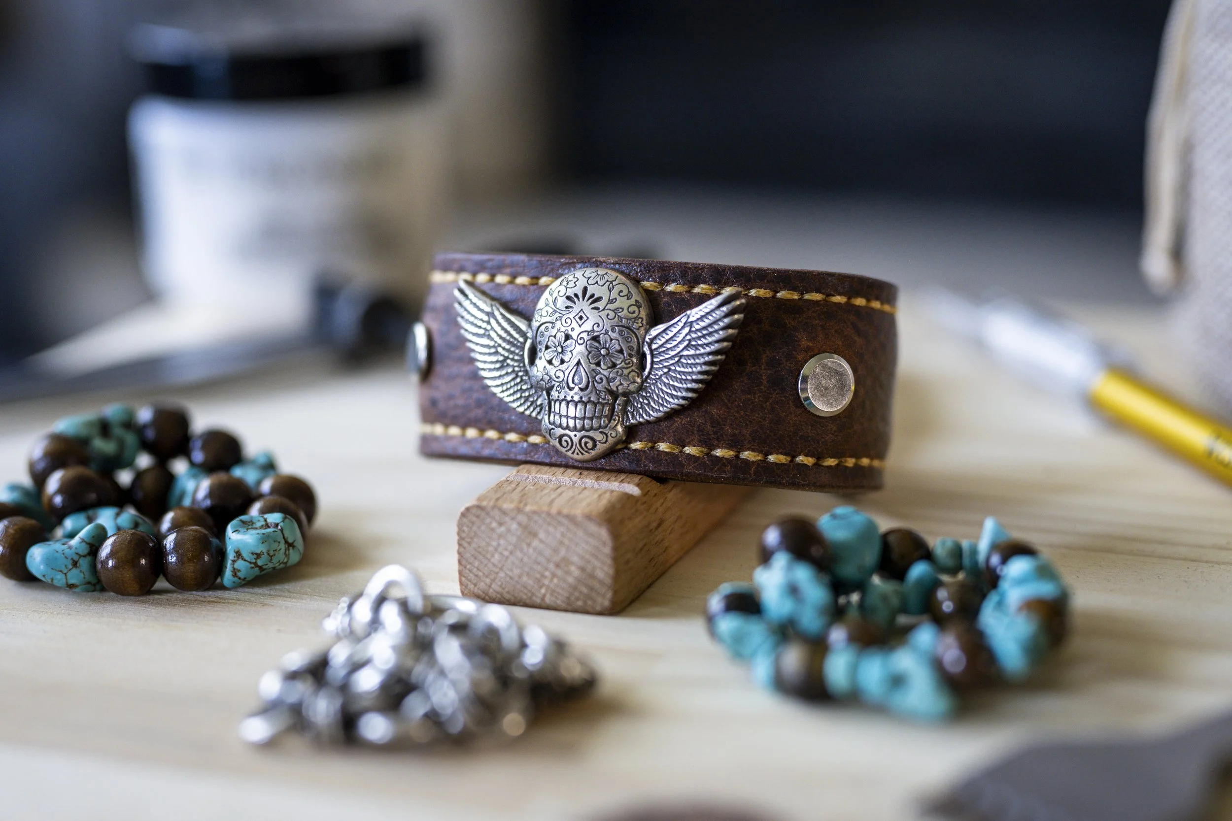 Close-up of a brown leather wristband with metal skull and wing embellishments, surrounded by turquoise and brown beaded jewelry on a light surface.
