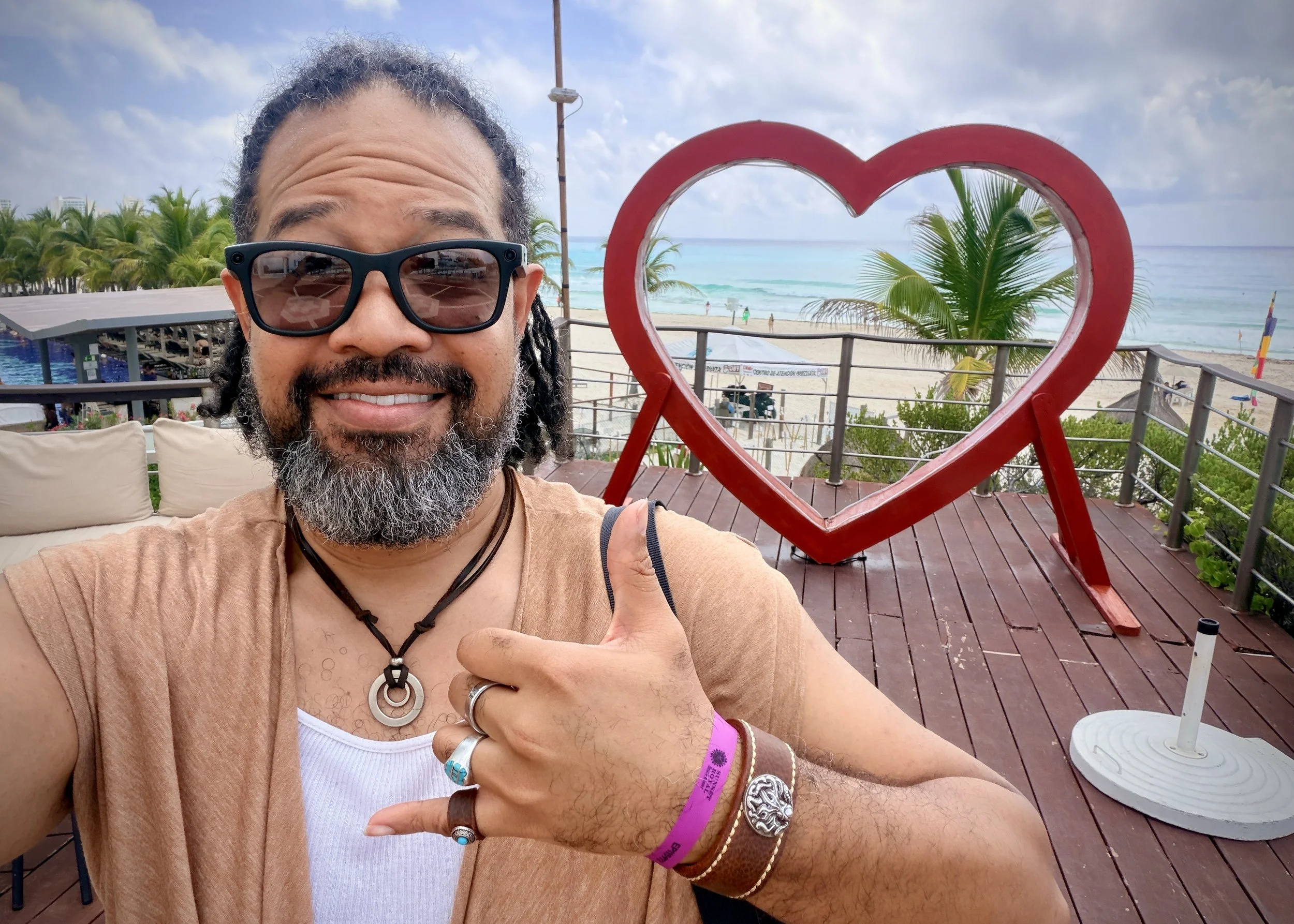 A smiling Black man wearing sunglasses gives a shaka beside a large red heart-shaped sculpture, with palm trees, ocean, and beach visible in the background.