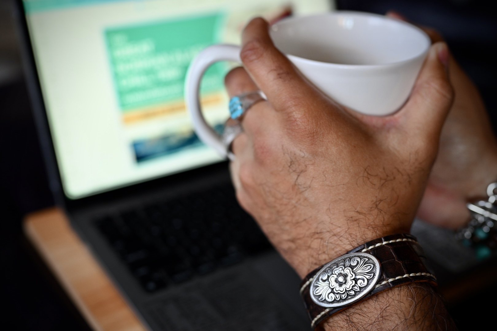 Close-up of a person's hand holding a white coffee mug over a laptop keyboard, with a webpage on the screen.