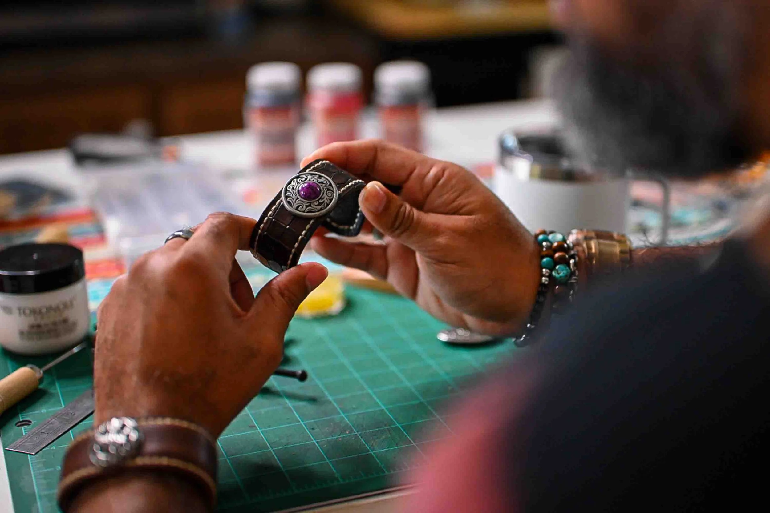 A person holding a silver and purple ring with intricate designs, surrounded by jewelry and crafting supplies on a green cutting mat.