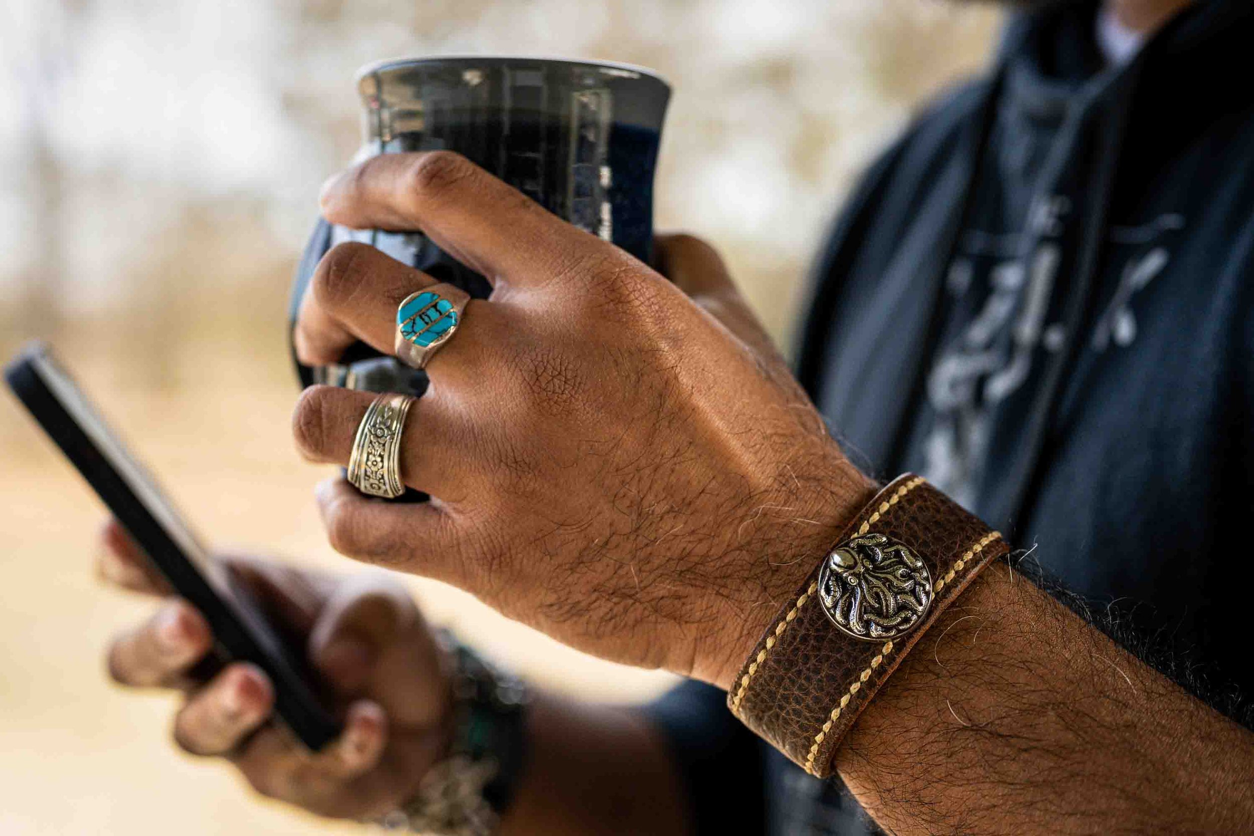 A hand wearing the Hilo leather cuff with an octopus concho and several turquoise rings holds a smartphone and takeaway coffee cup against a softly blurred outdoor background.