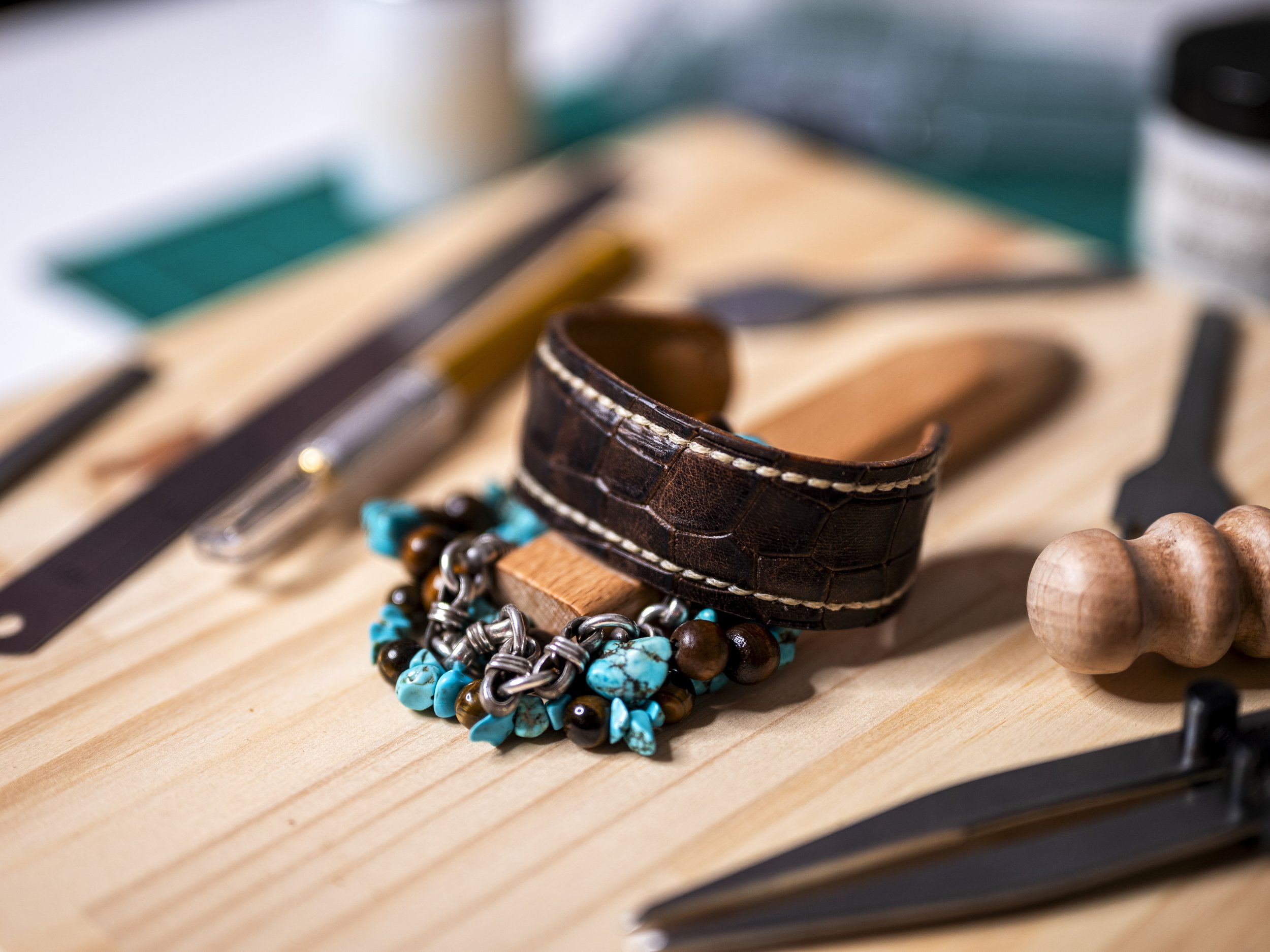 A handcrafted brown leather cuff sits among turquoise, brown, and metallic beads and leatherworking tools arranged on a wooden workbench.