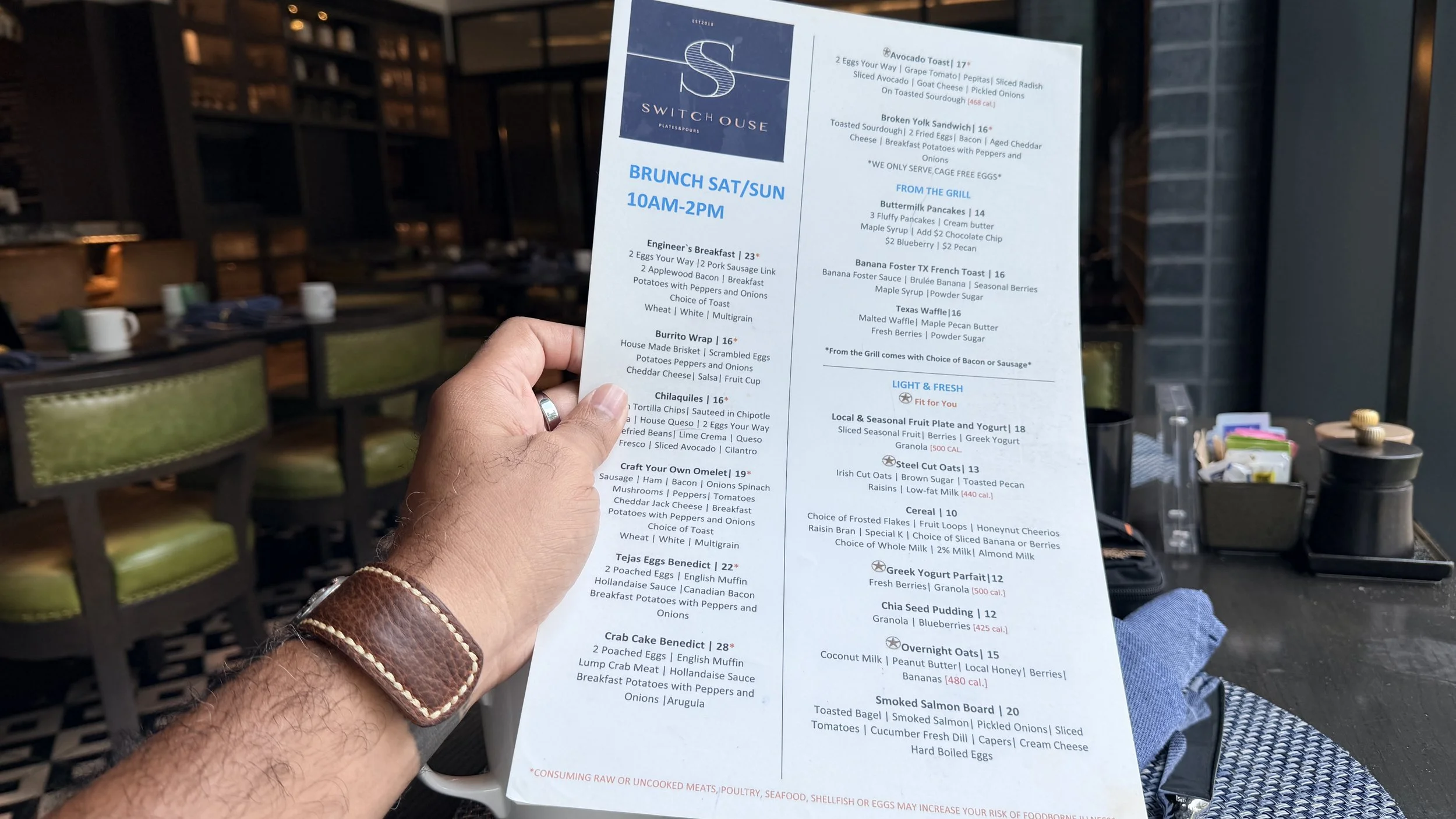 A Black man’s hand wearing a handcrafted brown leather cuff holds a brunch menu inside Switch House Plates & Pours, with the restaurant interior visible in the background.