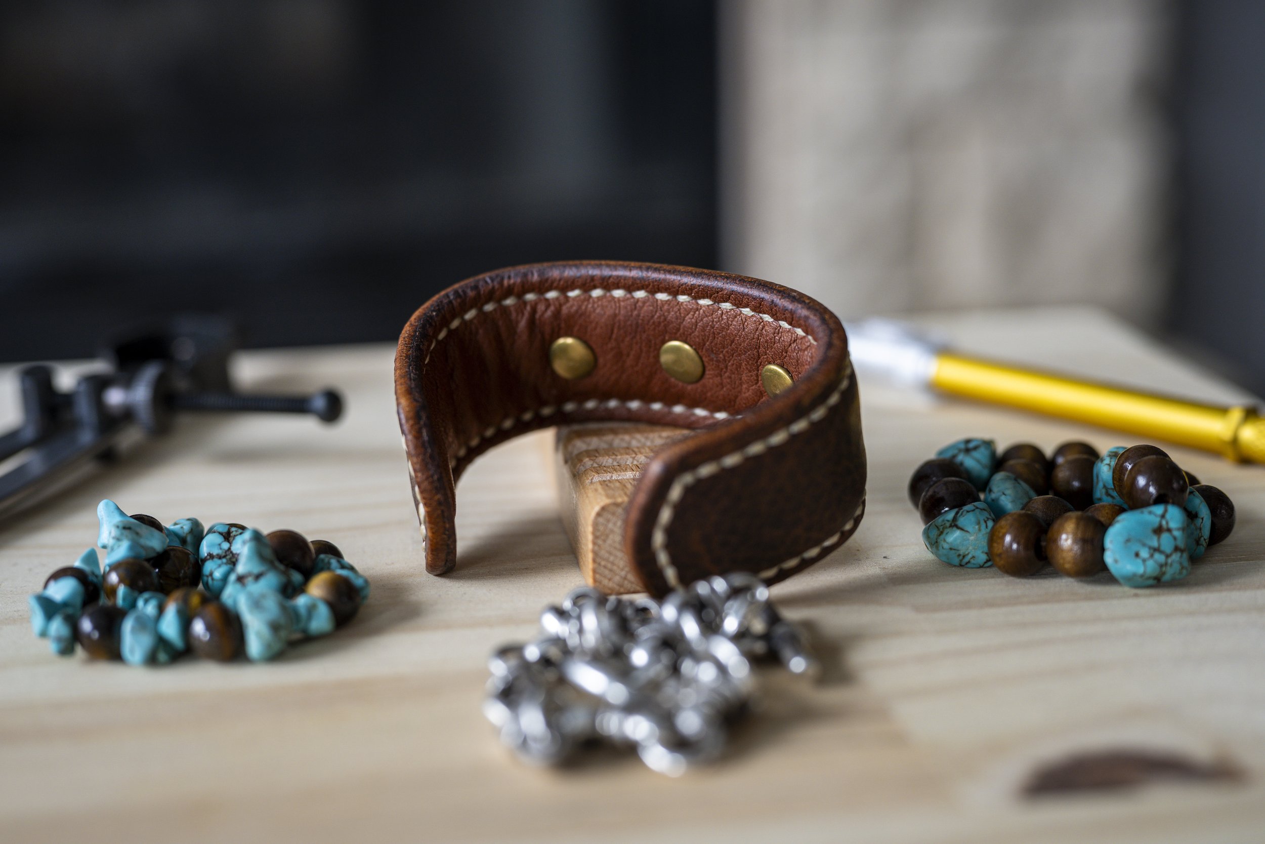 Rear view of the handcrafted Koa brown leather cuff, showing its brass-toned hardware and contrasting stitching, displayed among turquoise-colored beads and silver-toned materials on a wooden workshop surface.