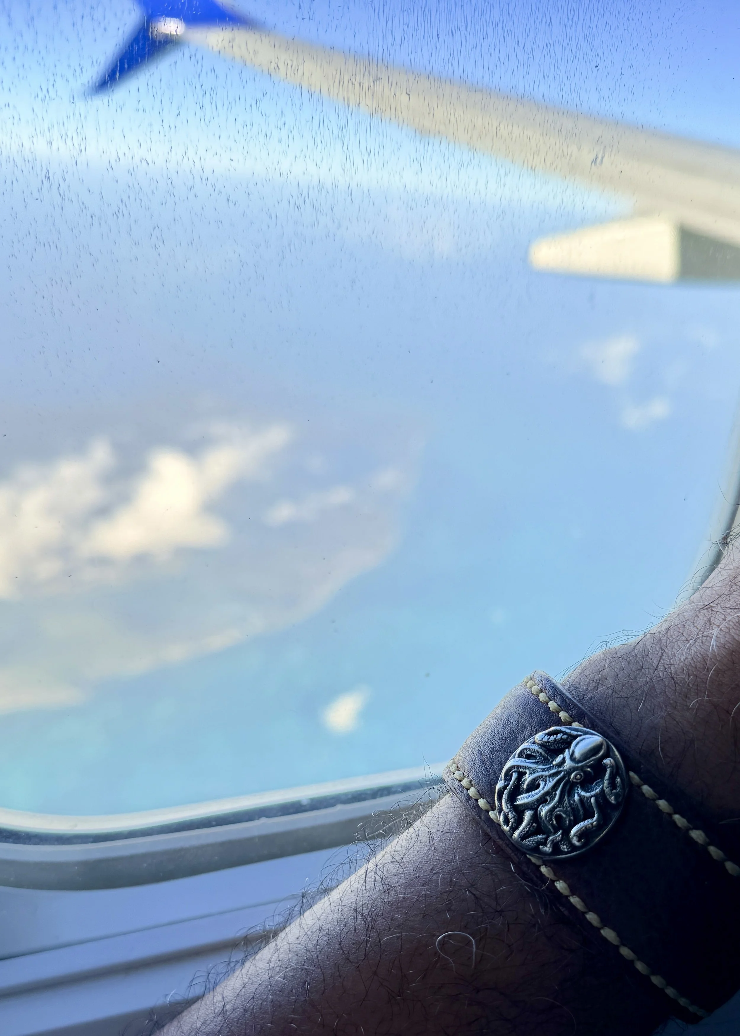 A Black man’s arm wearing the Hilo handcrafted brown leather cuff rests beside a rain-speckled airplane window, with the wing and clouds visible outside.